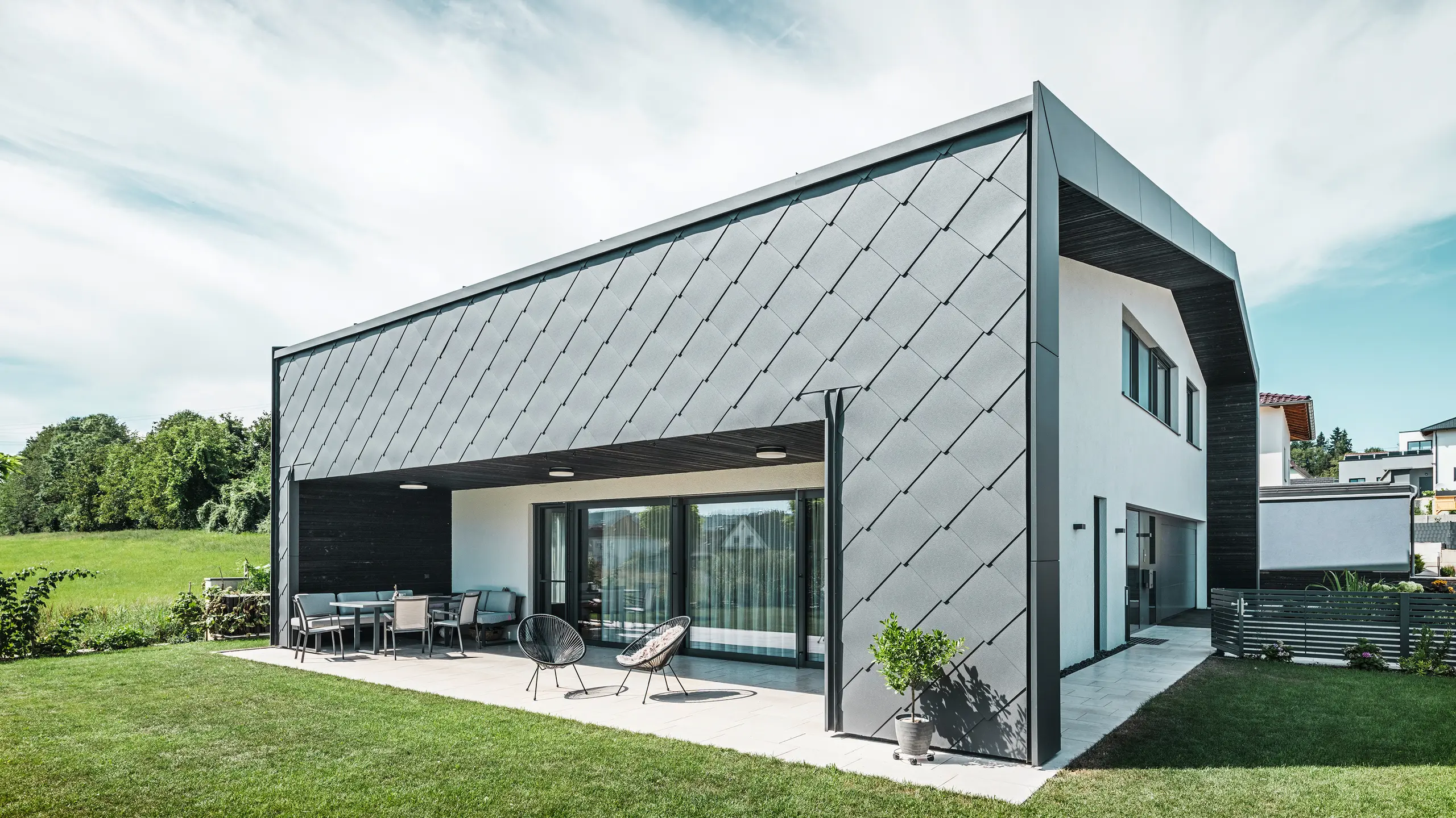 Upravená zahrada rodinného domu v Aschachu. Budova je stylově pokryta tmavě šedými fasádními šablonami PREFA Aluminium 44 × 44. Krytá terasa nabízí příjemné posezení s výhledem na svěží zelený trávník. Elegantní hliníkový obklad v kombinaci s bílými omítkami zajišťuje moderní a povětrnostním vlivům odolnou architekturu. Velké okna propouštějí do interiéru dostatek denního světla a vytvářejí otevřenou, světlou atmosféru.