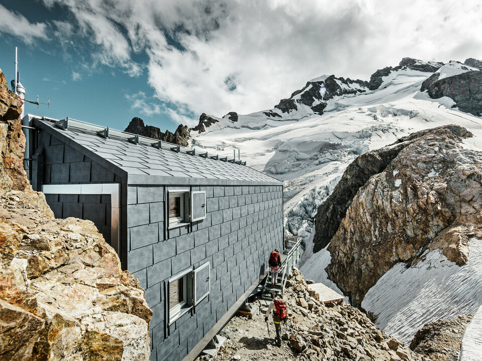 Seitliche Ansicht der Refuge de l'Aigle Berghütte mit steingrauen FX.12 Dach- und Fassadenpaneelen von PREFA, eingebettet in eine beeindruckende Gletscherlandschaft.