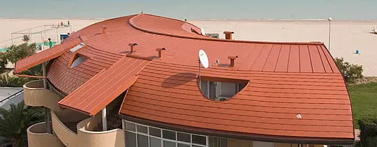 Zaoblená střecha souboru bytových domů, krytá PREFA falcovanými střešními šindeli a PREFA střešní krytinou Prefalz se stojatou drážkou v cihlově červené barvě.