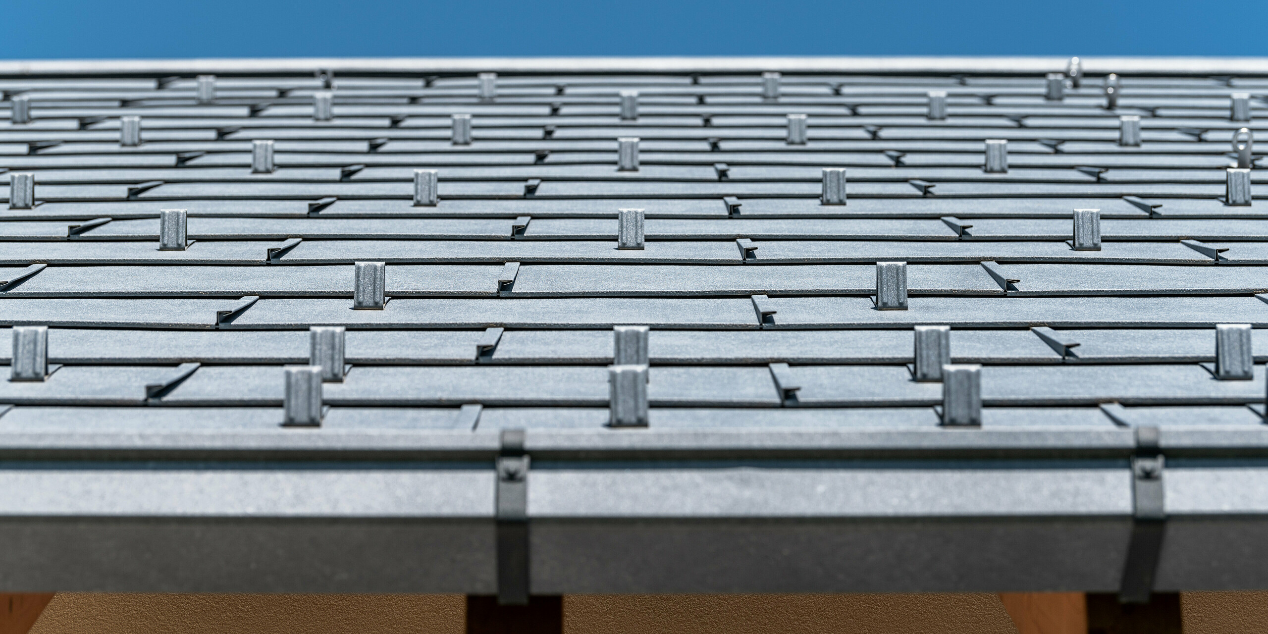 Detailní pohled na střechu PREFA pokrytou střešním panelem R.16 v barvě P.10 antracit. Na obrázku jsou precizně zpracované hliníkové střešní panely. Záběr vyzdvihuje výraznou strukturu a moderní design střechy, který kontrastuje s jasně modrou oblohou a podtrhuje inovativní a odolné řešení zastřešení PREFA.
