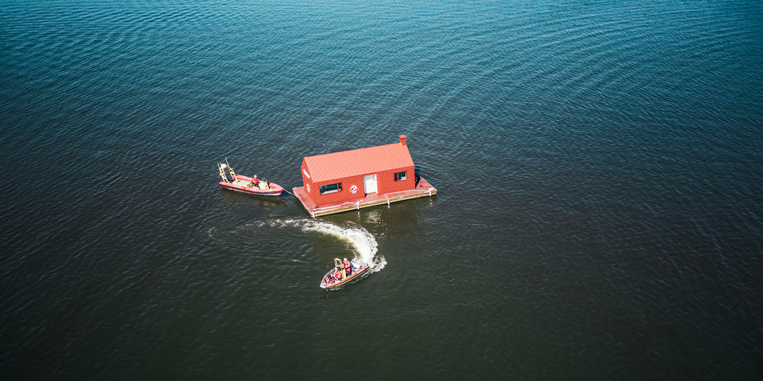 Pohled z ptačí perspektivy na poutavý dům námořní záchranné stanice na jezeře Hjälmaren ve Švédsku s tmavě červeným hliníkovým pláštěm vyrobeným z PREFA hliníkových šablon 29×29 tiše leží na vodě. Vycházející slunce za budovou zalévá scénu měkkým světlem, které se odráží ve stojaté vodě spolu s nádražním domem a záchrannými čluny.