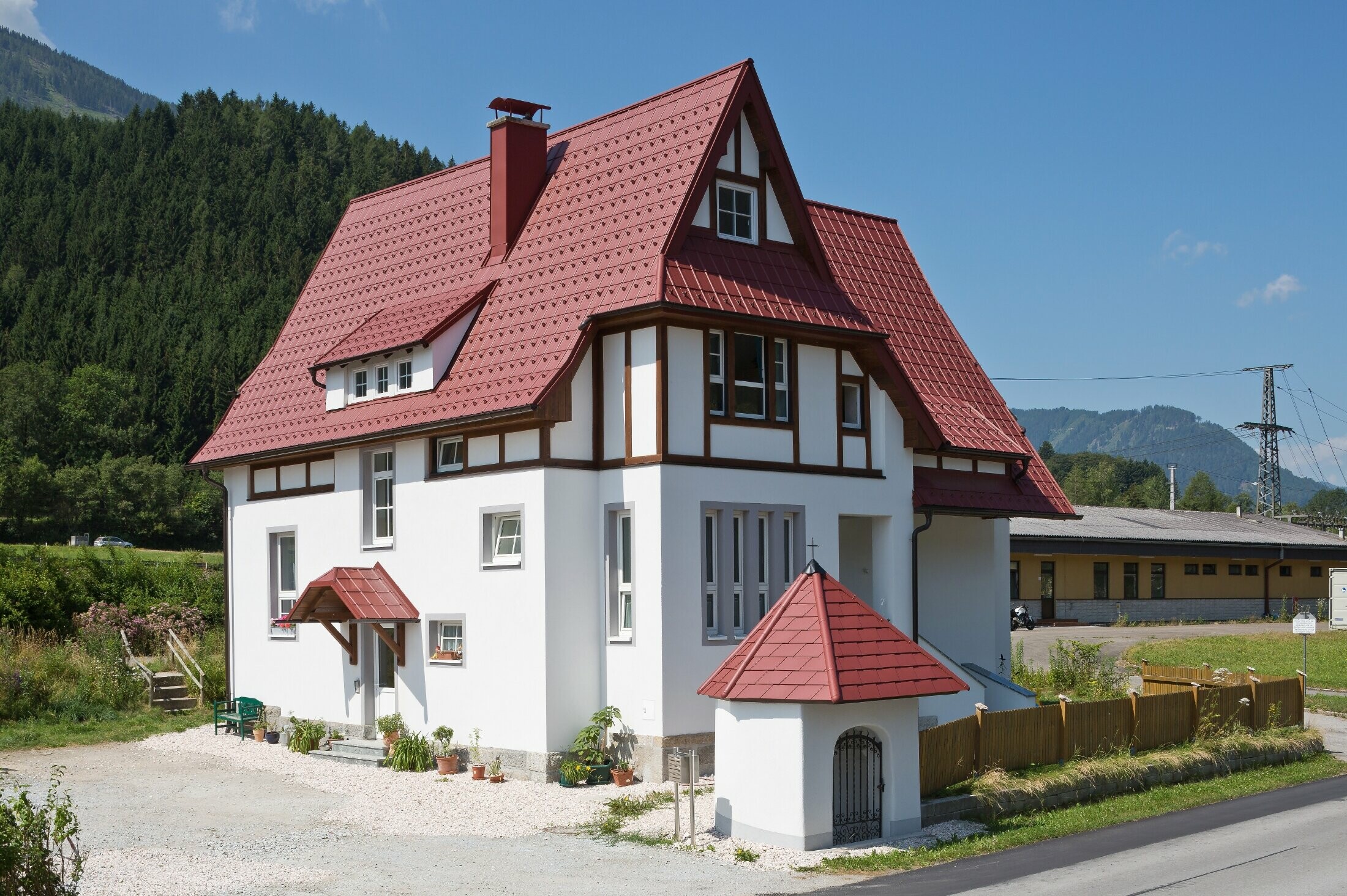 Verwinkeltes Steildach mit PREFA Dachplatte in P.10 Oxydrot mit vielen Details wie Gauben, Türmchen und Kamineinfassungen. Auch das Vordach und das Marterl vor dem Haus sind mit PREFA Blechdach eingedeckt.