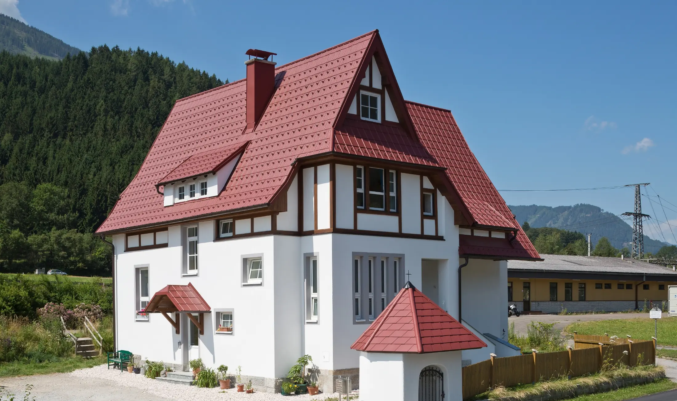 Šikmá střecha se falcovanou taškou PREFA v P.10 tmavě červené barvě s mnoha detaily, jako jsou vikýře, věžičky a komínová tělesa. Stříška a kaplička před domem jsou rovněž pokryty plechovou krytinou PREFA.