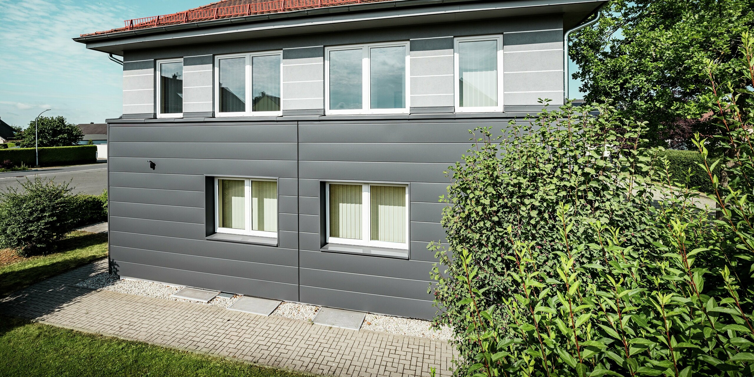 Das Bild zeigt die modernisierte Fassade der Kaminkehrerinnung in Bindlach, die mit PREFA Sidings in der eleganten Farbe P.10 Dunkelgrau verkleidet wurde. Die klare Struktur der Aluminium-Paneele schafft eine ansprechende, zeitgemäße Optik, während die robuste Oberfläche der Fassade hervorragenden Schutz vor Witterungseinflüssen bietet. Das Gebäude erhält durch die harmonische Kombination aus den dunkelgrauen Sidings und den grünen Pflanzen eine frische, lebendige Ausstrahlung. Dank der langlebigen, pflegeleichten Materialien bleibt die Fassade über Jahre hinweg in erstklassigem Zustand und sorgt für eine dauerhaft ästhetische Erscheinung.