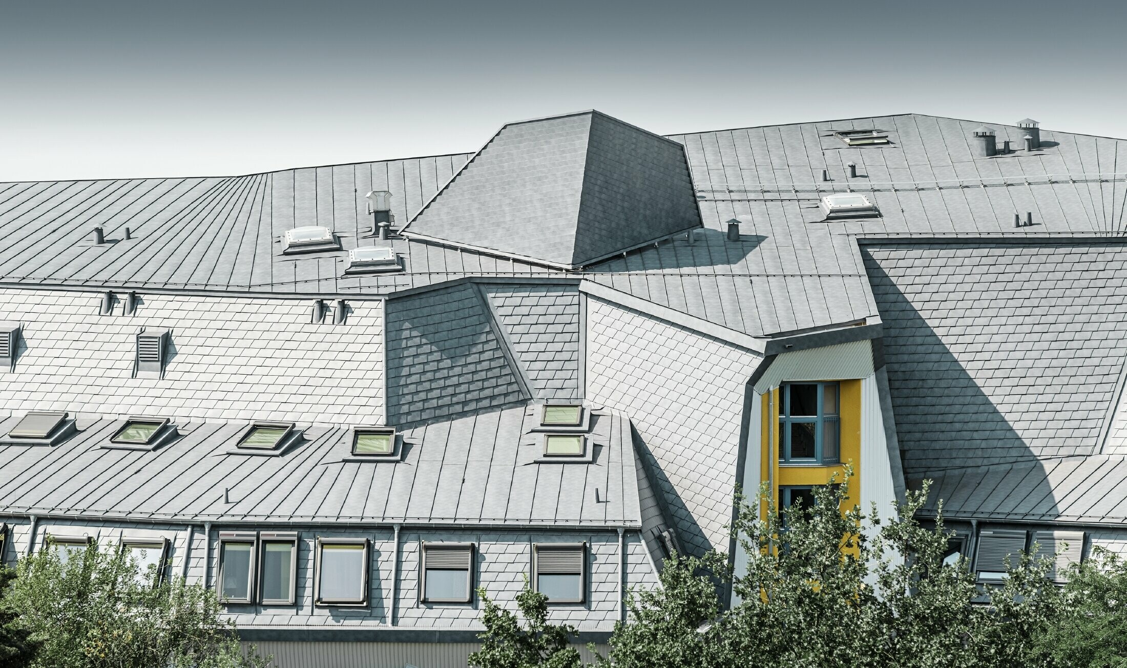 Členitá střecha s vikýři a klikatými rohy pečovatelského domu pokrytá falcovaným šindelem a svitkovým plechem PREFALZ v barvě P.10 břidlicové