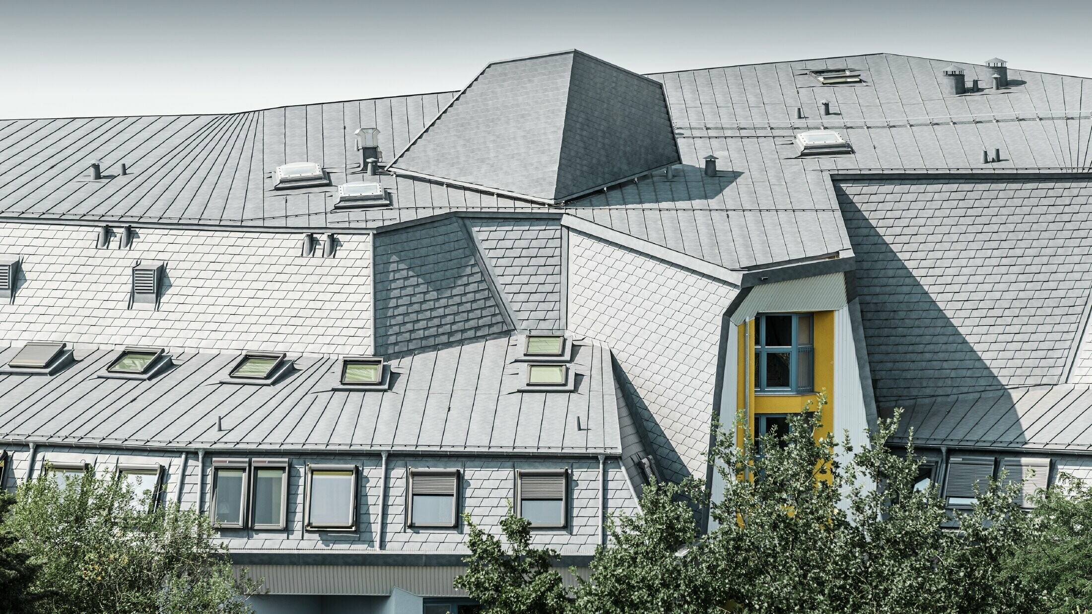 Členitá střecha s vikýři a klikatými rohy pečovatelského domu pokrytá falcovaným šindelem a svitkovým plechem PREFALZ v barvě P.10 břidlicové