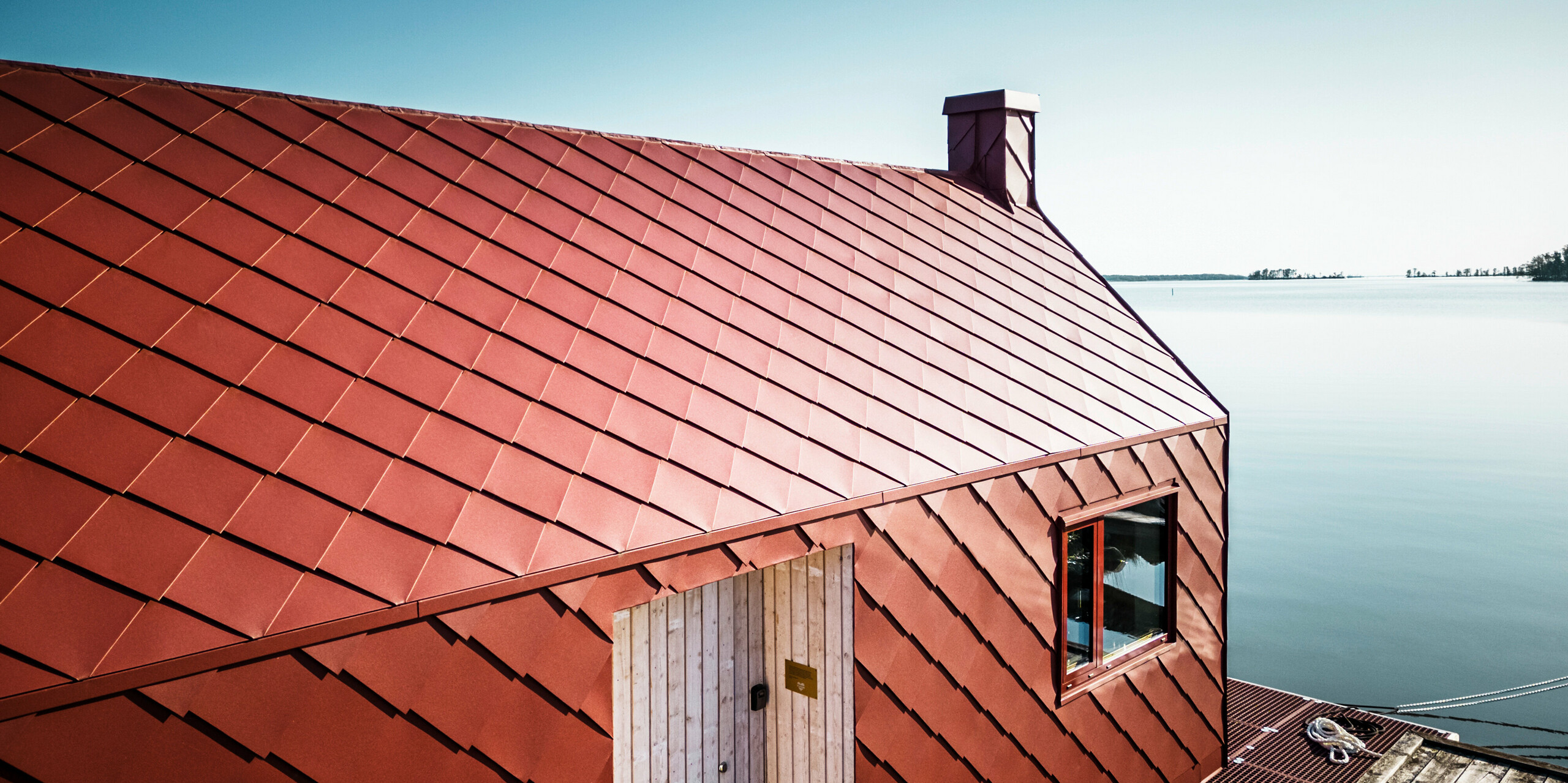 Architektonický detailní záběr plovoucí stanice vodních záchranné služby na jezeře Hjälmaren s velkolepým hliníkovým pláštěm PREFA vyrobeným z falcovaných a fasádních šablon 29×29 v barvě P.10 tmavě červené. Precizně uspořádané šablony na střeše a fasádě odrážejí odolnost PREFA a dodávají budově moderní a příjemný vzhled. Plovoucí nádražní dům musí odolávat ledu přibližně polovinu roku a je optimalizován pro vysoké zatížení způsobené větrem a vlnami.