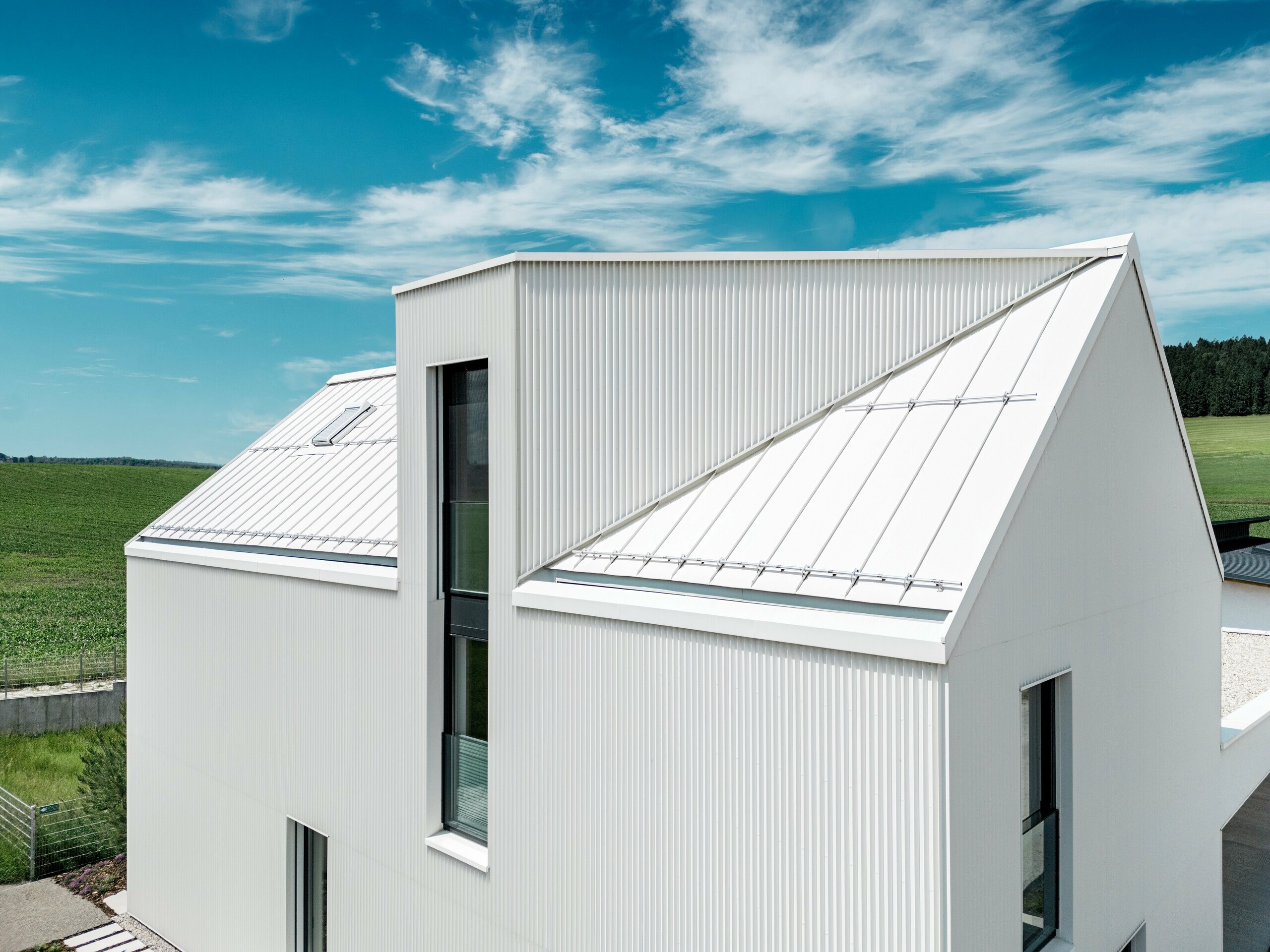 Obrázek ukazuje strmou, výraznou sedlovou střechu tohoto rodinného domu ve Freistadtu, obloženou materiálem PREFALZ v čistě bílé barvě. Precizně skládaná fasáda a střecha jsou bezproblémově integrovány a vytvářejí elegantní a čistý estetický dojem. PREFALZ použitý na střeše i fasádě zajišťuje nejen estetickou trvanlivost, ale také vysokou odolnost vůči povětrnostním vlivům. To budově propůjčuje dlouhodobou životnost a snadnou údržbu. Souhra moderního designu a funkčních materiálů vytváří harmonickou, moderní architekturu, která je praktická i vizuálně přitažlivá. Obzvláště poutavé je vikýřové okno se svým šikmým designem a dvoupatrovou přístavbou.