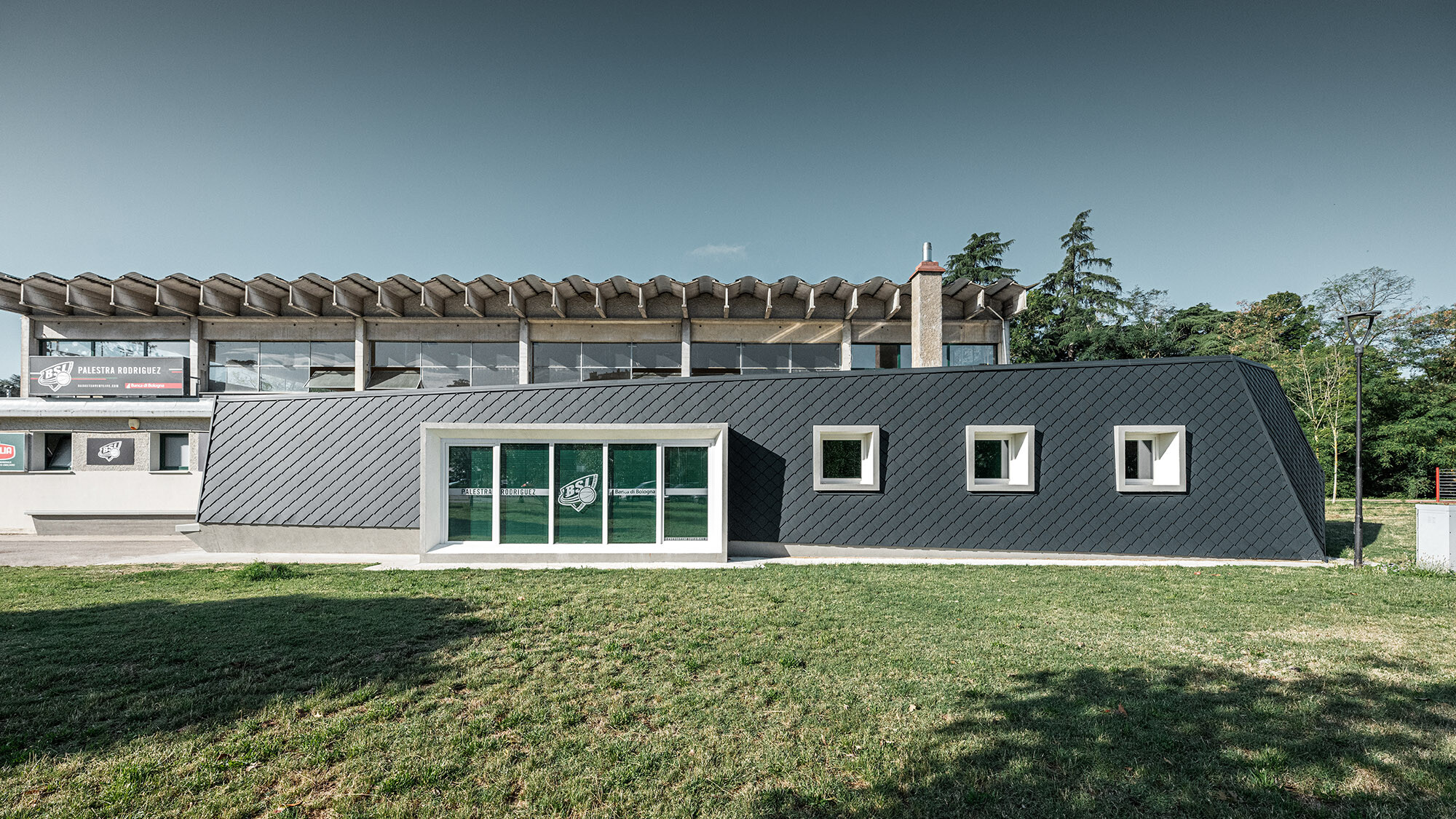 Pohled na moderní tělocvičnu v italské Bologni s fasádou z antracitových hliníkových šablon od PREFA ve formátu 20 x 20 cm, na zalesněném pozadí a pod jasnou oblohou.