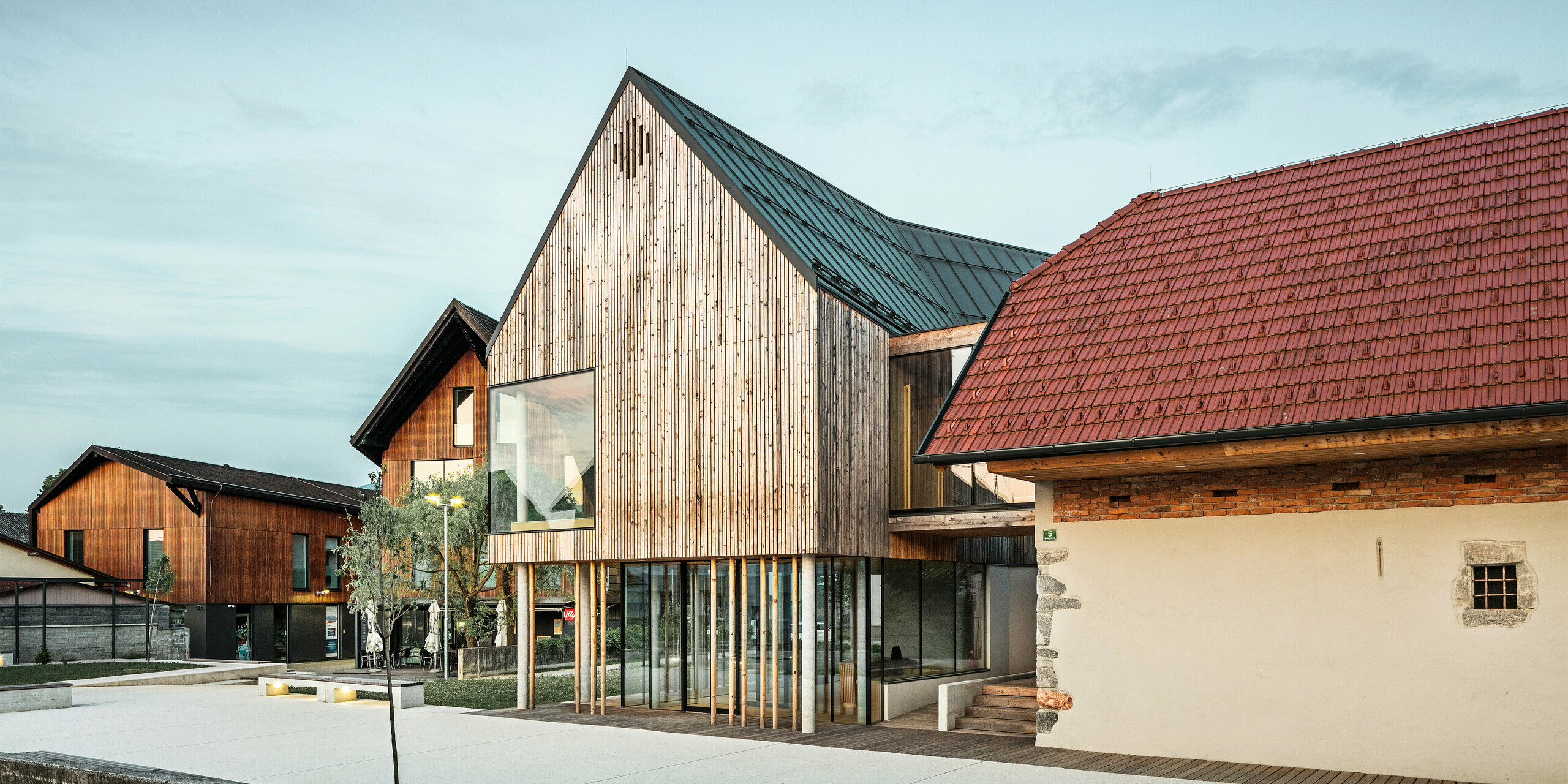 Soumrakový pohled na návštěvnické centrum ve slovinském Ig s nápadnou střechou PREFALZ v barvě P.10 tmavě šedá, která výrazně vyčnívá z okolní tradiční červené taškové střešní krytiny. Spojení starého a nového, reprezentované přilehlou stěnou s malým oknem a moderní dřevěnou konstrukcí s velkými prosklenými plochami, svědčí o promyšlené symbióze regionální tradice a současné architektury. Stylové osvětlení podtrhuje lákavou atmosféru vstupního prostoru, jak je prezentováno v PREFARENZEN book 2023.
