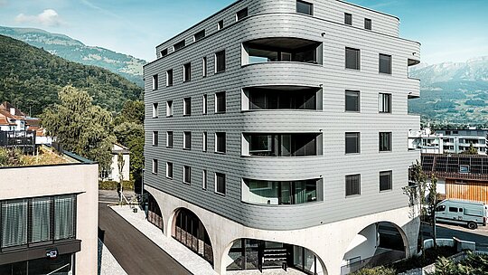 Vnější pohled na Bernerhaus v Buchsu s jeho výrazným trojúhelníkovým tvarem a jemně zaoblenými balkony. Fasáda, obložená PREFA fasádními šindeli v barvě P.10 světle šedé, obaluje budovu jako šupinovitý hliníkový plášť a vytváří precizní a živoucí fasádu. Nad masivním betonovým základem s elegantními arkádami působí obzvláště silně lehká, povětrnostním vlivům odolná hliníková fasáda PREFA. Odolná, nenáročná na údržbu a rozměrově stabilní udává moderní architektonický tón na pozadí alpských hor.