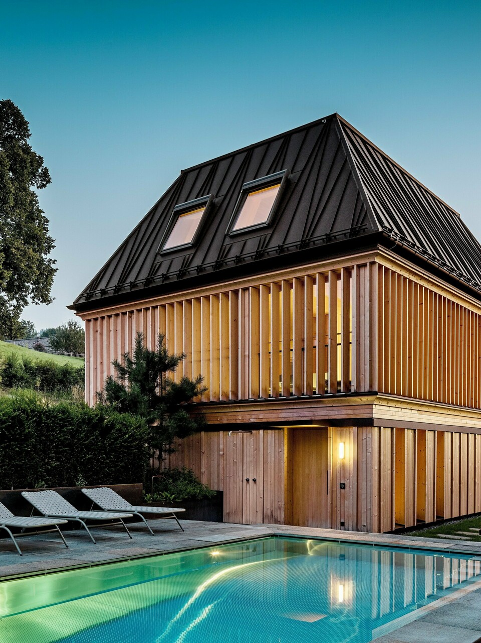 „Lázně“ hotelu Hirschen ve Schwarzenbergu za soumraku s výhledem na osvětlený venkovní bazén v popředí. Budova zaujme moderní valbovou střechou z PREFALZu v oříškově hnědé barvě P.10 s precizně provedenými, částečně zkosenými stojatými drážkami. Dvě střešní okna zajišťují přirozené světlo uvnitř. Fasáda se skládá z vertikálních dřevěných lamel od podlahy až ke stropu, které dodávají budově strukturu a lehkost. Ve spodní části převládá přírodní dřevo. Obklopen upravenou zelení a zasazený do zvlněných kopců Bregenzerwaldu, vytváří harmonickou směs architektury, přírody a relaxace.