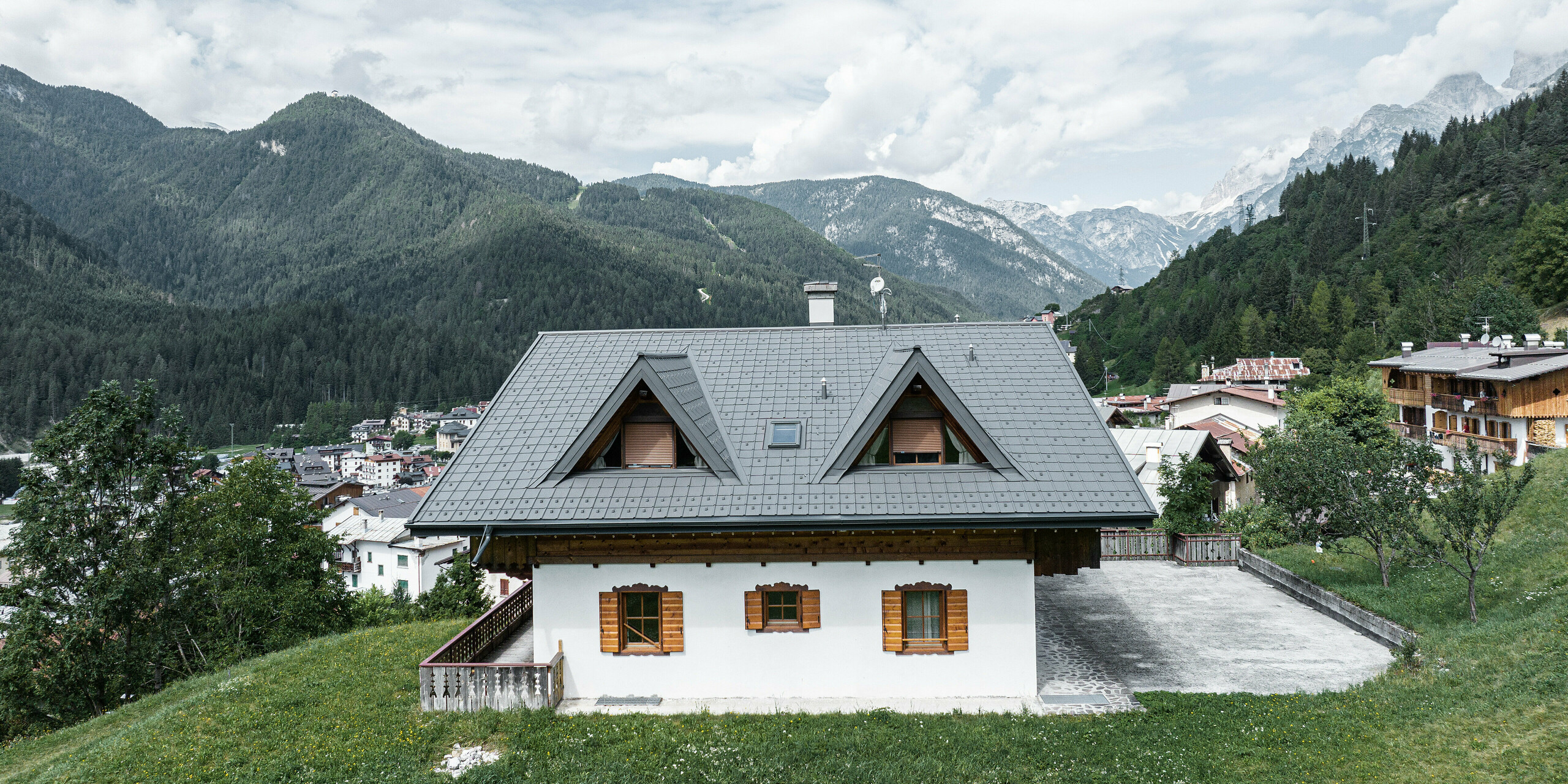 Zadní strana horské chaty v Auronzo di Cadore v Itálii ukazuje čerstvě nainstalované falcované tašky PREFA ve světle šedé barvě. Dům stojí na zelené louce s majestátními Dolomity v pozadí. Dva špičaté vikýře dokonale zapadají do horského pozadí v pozadí. Pečlivá rekonstrukce střechy podtrhuje tradiční kouzlo budovy, zatímco moderní prvky ji mísí s alpskou krajinou.