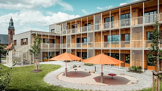 Zadní část školky v Alzenau odhaluje chráněný dvůr s přilehlým hřištěm. Dva oranžové slunečníky dodávají barevné paprsky nad kruhovým pískovištěm. V pozadí se obytné křídlo, postavené z udržitelného masivního dřeva, rozšiřuje o jemnou, vyčnívající kovovou balkonovou a schodišťovou konstrukci. Vlevo je viditelná hliníková fasáda obložená bronzovými fasádními šablonami PREFA 44 x 44 – robustní a esteticky příjemná forma ochrany před povětrnostními vlivy. Kombinace hliníku, dřeva a oceli symbolizuje inteligentní integraci funkčnosti, udržitelnosti a architektonické kvality. Pohled na kostel v pozadí ukotvuje projekt v zavedené městské krajině Alzenau.
