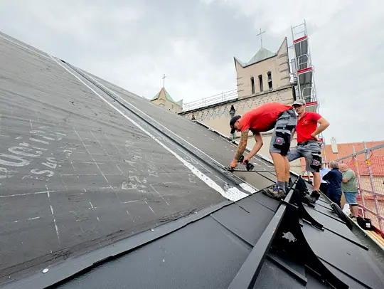 Obrázek ukazuje instalaci fotovoltaických systémů integrovaných do střechy kostela v Bavorsku. V popředí jsou jasně viditelné solární střešní panely, úhledně poskládané v řadách. Panely mají tmavý, lesklý povrch. V pozadí je vidět část střechy kostela a jeho charakteristické prvky, jako je věž a kříž. Na střeše jsou dva dělníci: jeden provádí instalační práce, zatímco druhý panely kontroluje.