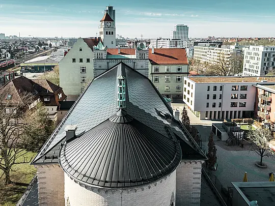 Das Bild zeigt das markante, rund gewölbte Dach auf der Rückseite der St. Karl Borromäus Kirche in Nürnberg. Die Gebäuderundung wurde mit einer Stehfalzdeckung aus schwarzem Aluminium eingedeckt. Das Satteldach, das den Hauptteil des Gebäudes schützt, wurde mit schwarzen PREFA Solardachplatten eingedeckt. Die innovativen Solardachplatten fungieren als optisches Highlight und liefern gleichzeitig Strom aus erneuerbarer Energie. Der Blick auf das Dach und die zentral positionierte Turmspitze verdeutlicht die harmonische Integration der modernen Solartechnologie in die historische Architektur. Im Hintergrund sind sowohl traditionelle als auch moderne Gebäude zu sehen, was die gelungene Verbindung von Denkmalschutz und nachhaltigem Bauen unterstreicht.