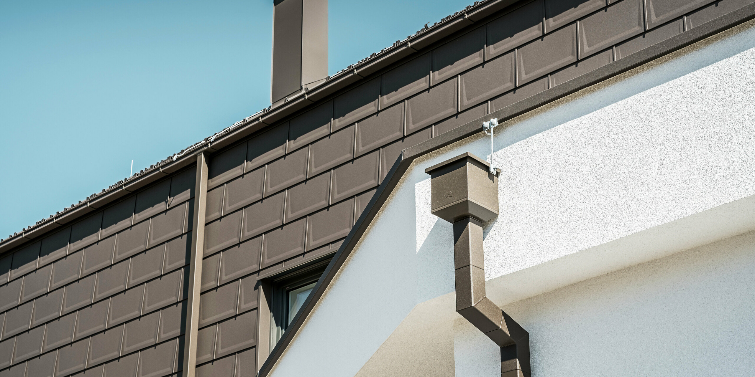 Detailní pohled na hranu střechy nebo fasádu rodinného domu v rakouském Neukirchenu, který je vybaven střešními panely PREFA R.16 v P.10 hnědé. Jak střešní panely, které v této budově fungují jako střešní a fasádní obklady, tak střešní odvodňovací systém, sestávající ze čtyřhranné trubky, sběrného boxu na vodu a boxového okapu, jsou ve stejné barvě a nabízejí konzistentní, vysoce kvalitní Dokončit. Použití výrobků PREFA dodává nemovitosti moderní vzhled, zatímco estetické začlenění odvodňovacích prvků podtrhuje funkční eleganci designu.
