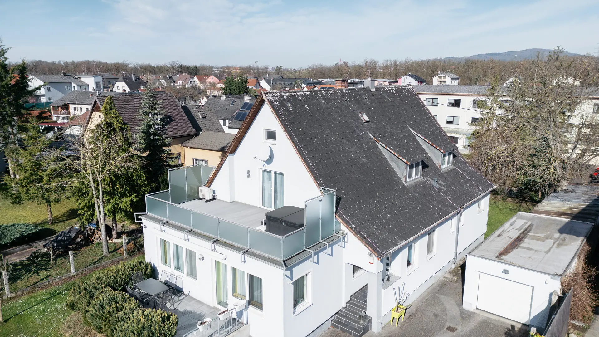 Pohled z ptačí perspektivy na budovu se starou eternitovou střechou pokrytou nečistotami a lišejníky. Na střeše nejsou vidět žádné solární panely. Balkon v popředí má skleněné zábradlí a nachází se v horním patře budovy.