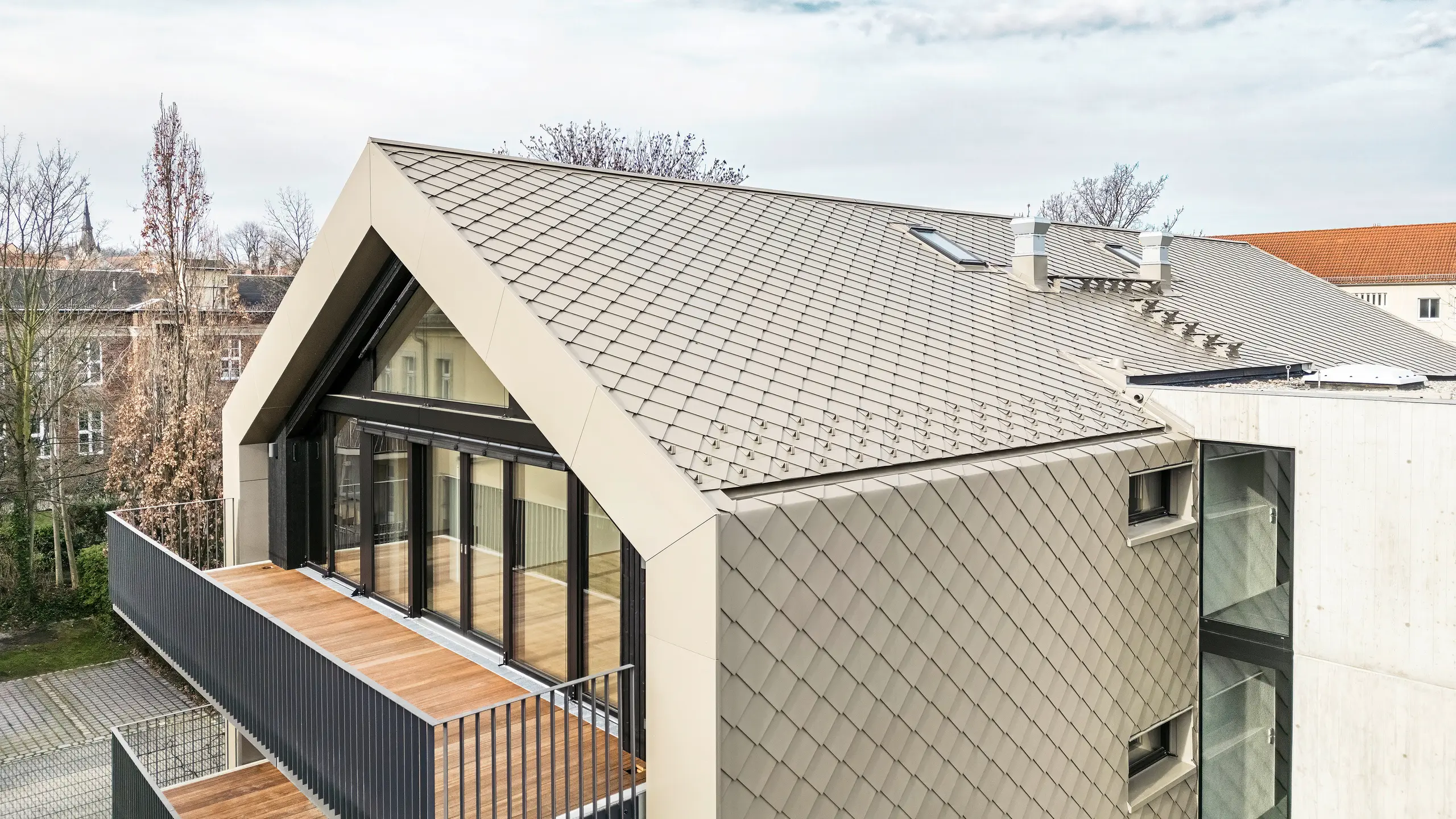 Pohled z ptačí perspektivy na moderní bytový dům na Chemnitzer Straße (Drážďany) s pláštěm falcovaných a fasádních šablon PREFA 29 × 29 v bronzu. Obrázek ukazuje elegantní střešní konstrukci, velká prosklená čela a balkony i harmonické začlenění budovy do okolí. Vysoce kvalitní materiály podtrhují dlouhodobou architekturu budovy.