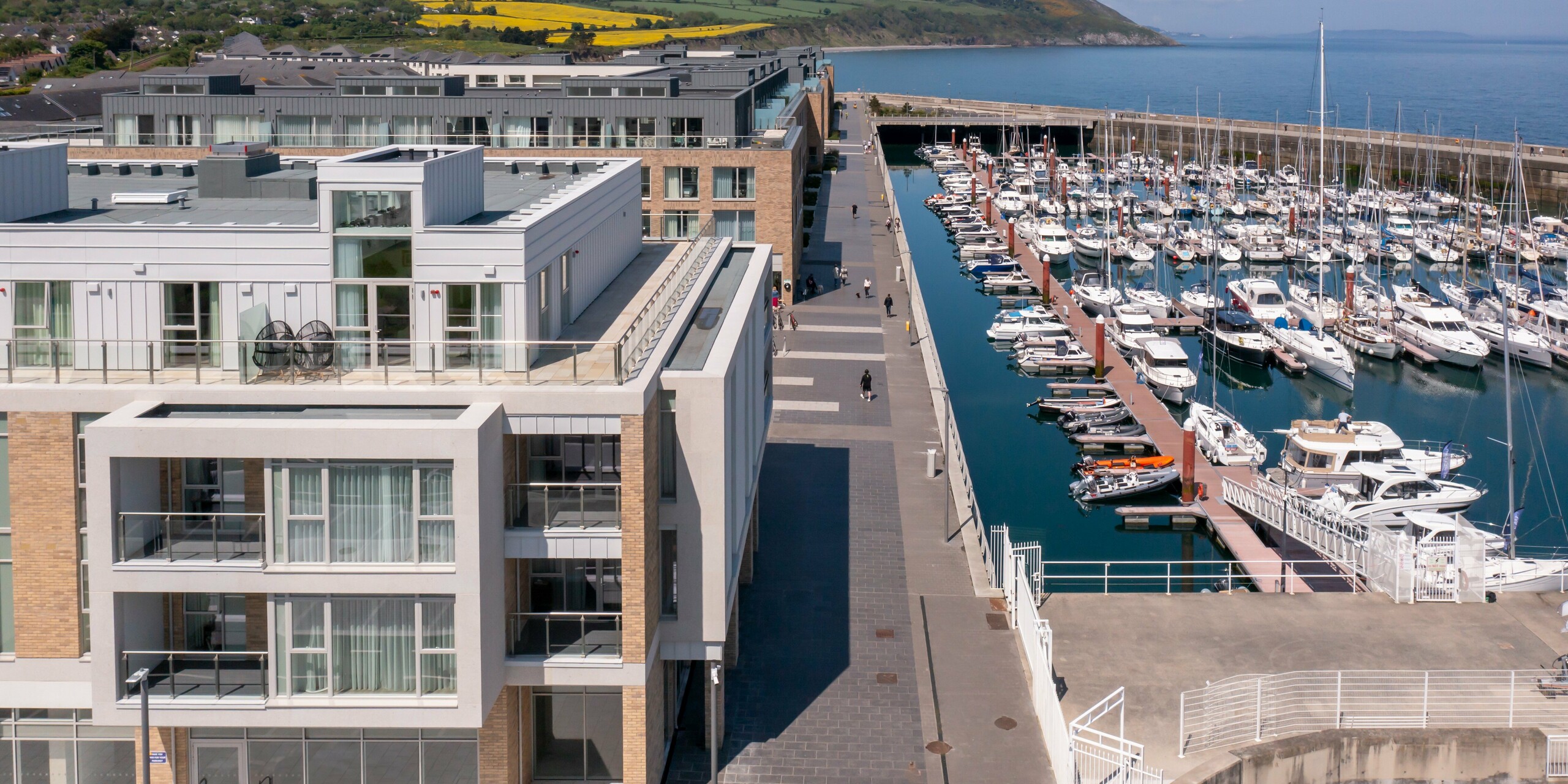 Letecký pohled na Marina Village Greystones ve Wicklow, Irsku. Moderní rezidenční zástavba přímo u přístavu. Fasáda rezidenčního komplexu je provedena z cca 2000 m² robustního PREFALZU v barvách P.10 prefa bílá a P.10 světle šedá. Elegantní architektura s prostornými prosklenými průčelími a balkony, obklopená malebnou pobřežní krajinou. Vysoce kvalitní hliníkové opláštění od PREFA nabízí trvalou ochranu a nadčasovou estetiku.