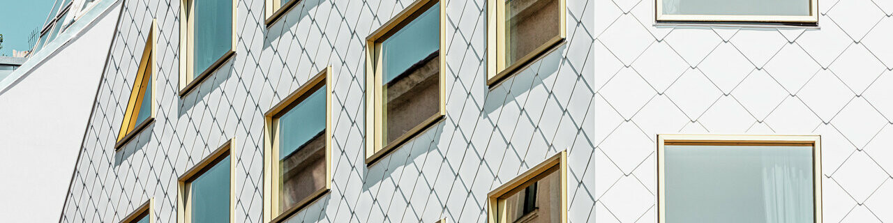 Moderní čtyřhvězdičkový hotel ve Vídni, pokrytý produkty PREFA: falcované a fasádní šablony  44 × 44 v barvě P.10 čistě bílé. Fasáda má kvádrovou strukturu s množstvím zrcadlově zasklených oken uspořádaných do ostrého rastru, odrážejícího pohyby okolí.