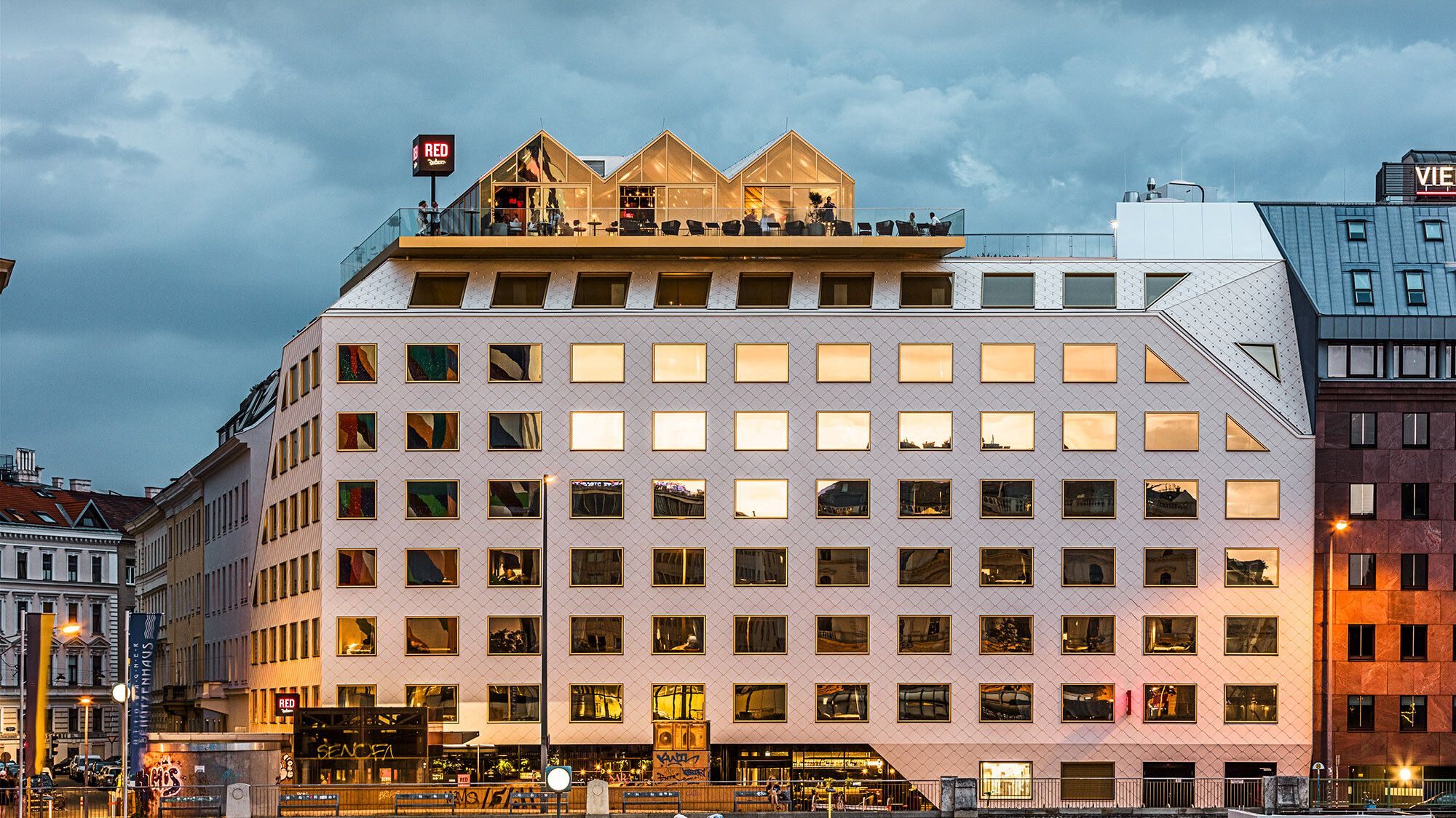 Večerní záběr hotelu THE ROCK Radisson RED Vienna a jeho živého okolí; v jeho oknech se odrážejí budovy naproti.