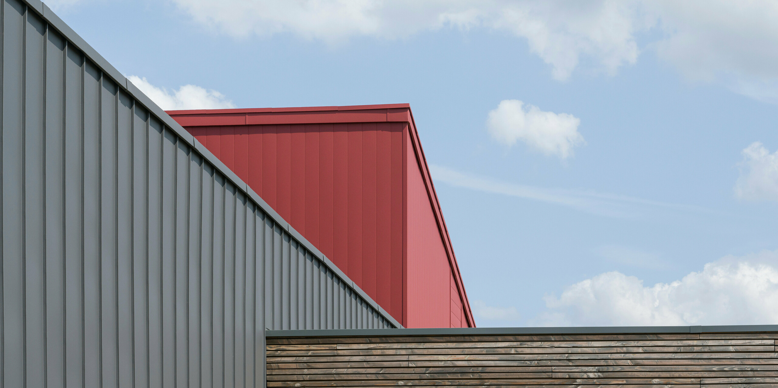 Detailní záběr na design střechy a fasády moderní budovy. V popředí je stěna s horizontálním dřevěným obkladem, který má přirozenou texturu. Za ní se tyčí tmavě šedá fasáda PREFALZ s vertikálními pásy, která vyzařuje industriální elegancí. Nad touto fasádou je část jasně červené hliníkové střechy a fasádního systému, která nabízí výrazný barevný kontrast a podtrhuje moderní architekturu.