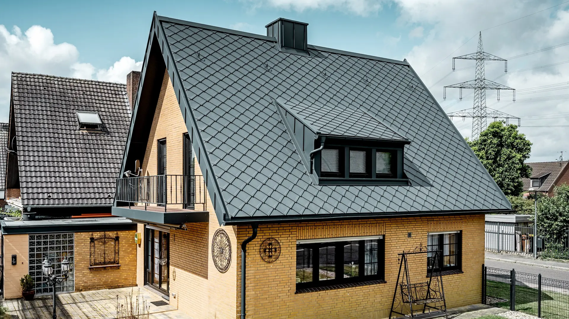 Rodinný dům ze žlutých pálených cihel po renovaci střechy s PREFA falcovanou šablonou 29 × 29 v tmavě šedé barvě; nově pokrytá, strmá sedlová střecha s kosočtvercovým vzorem, integrovanou vikýřem a komínem, terasou a zahradou v popředí.