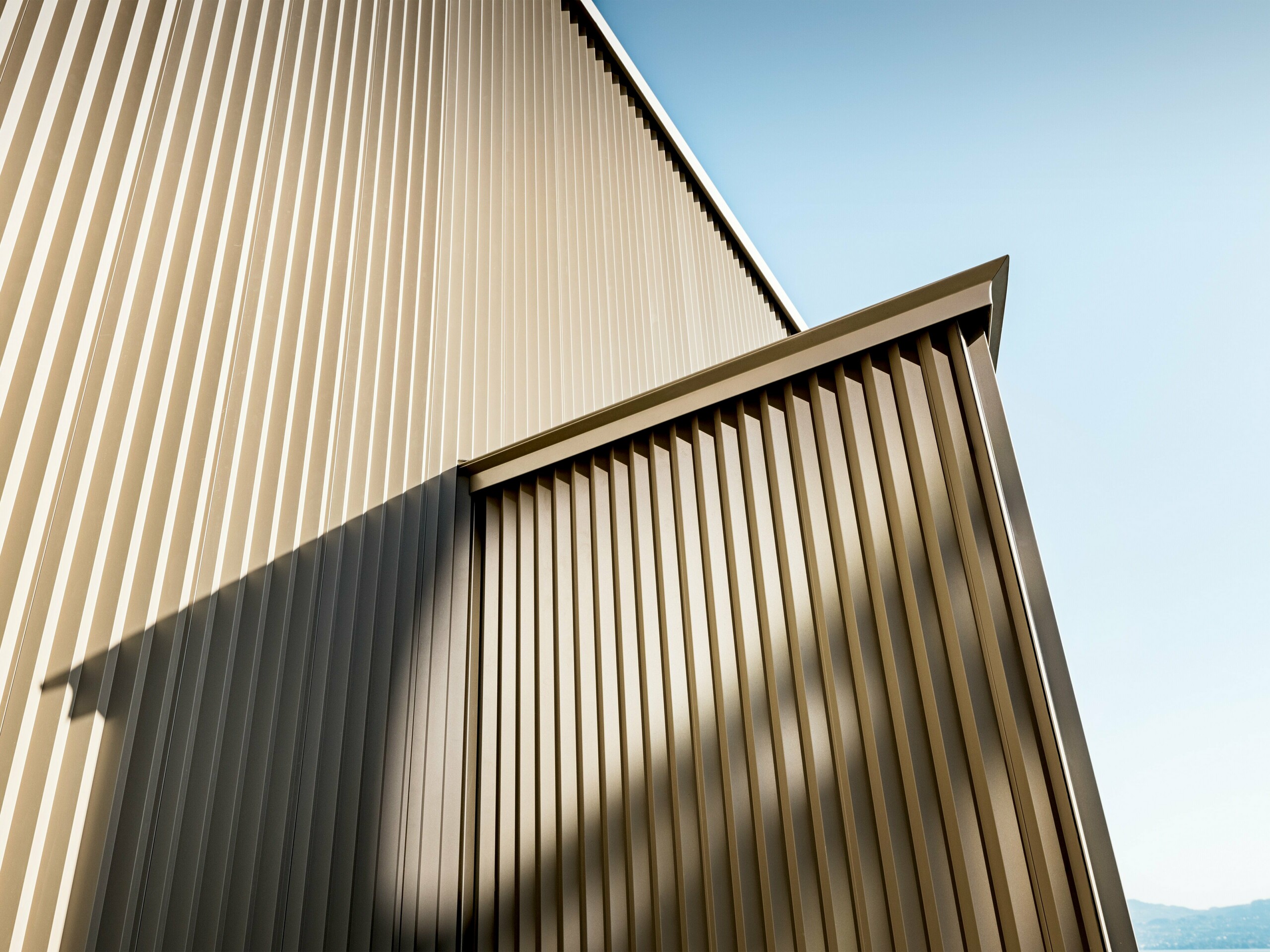 Elegantní hliníková fasáda PREFA s bronzovou povrchovou úpravou zdobí obytný dům v St. Gingolph, který navrhla architektka Sophie Morard. Svisle zoubkované, na míru vyrobené profily vytvářejí podmanivou hru světla, stínu a textury, což fasádě dodává hloubku a dynamiku. Vysoce kvalitní práškově lakovaný povrch zajišťuje dlouhodobou ochranu a stylovou estetiku – ideální pro sofistikované rezidenční projekty. Třpytivý bronzový design harmonicky splývá s alpským prostředím a proměňuje malý dům v architektonický vrchol s výrazným působením.