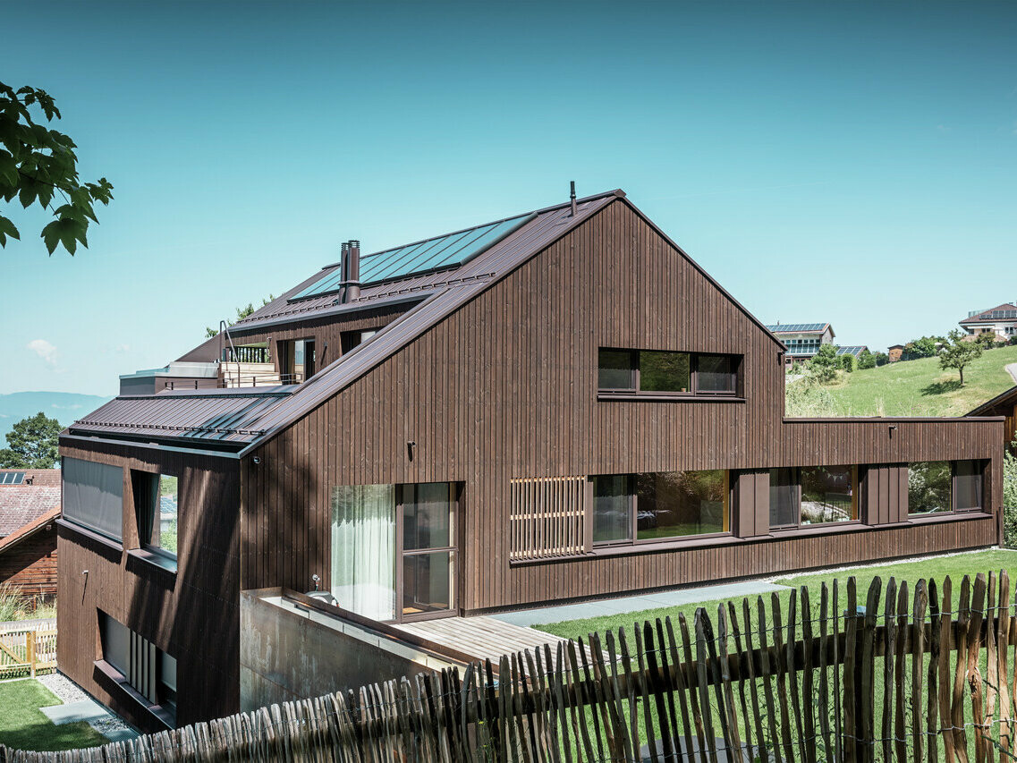 Moderní činžovní dům v rakouském Vorarlbersku se sedlovou střechou pokrytou Prefalzem v ořechově hnědé barvě. Dřevěná fasáda budovy harmonicky zapadá do venkovského prostředí, zatímco velká okna dodávají moderní akcenty.