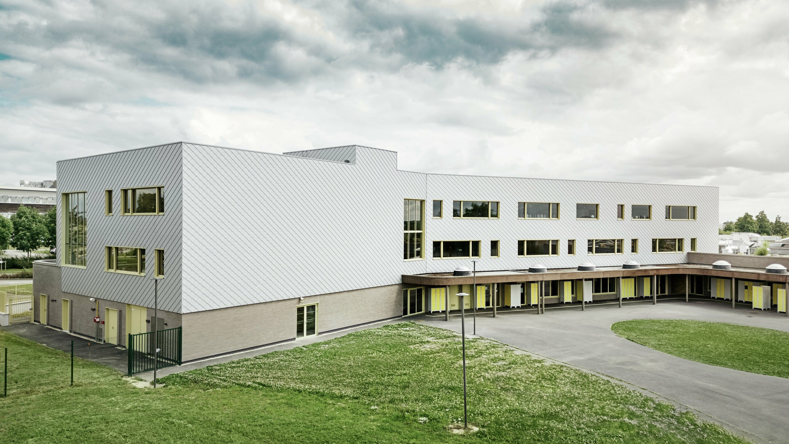Das Bild zeigt die Rückseite des Collège Léontine Dolivet. Im Vordergrund befindet sich ein großzügiger, asphaltierter Schulhof mit klaren Linien und offenen Flächen, die für Pausen und Freizeitaktivitäten genutzt werden können. Einige Grünflächen lockern die Umgebung auf. Im Hintergrund erhebt sich das moderne Schulgebäude mit seiner markanten Aluminiumfassade aus silbermetallic PREFA Wandrauten, die das Licht reflektieren und einen dynamischen Effekt erzeugen. Mehrere große Fenster sorgen für Helligkeit im Innenbereich. Die Szene ist bei Tageslicht aufgenommen, mit leicht bewölktem Himmel, was die metallische Oberfläche besonders betont.