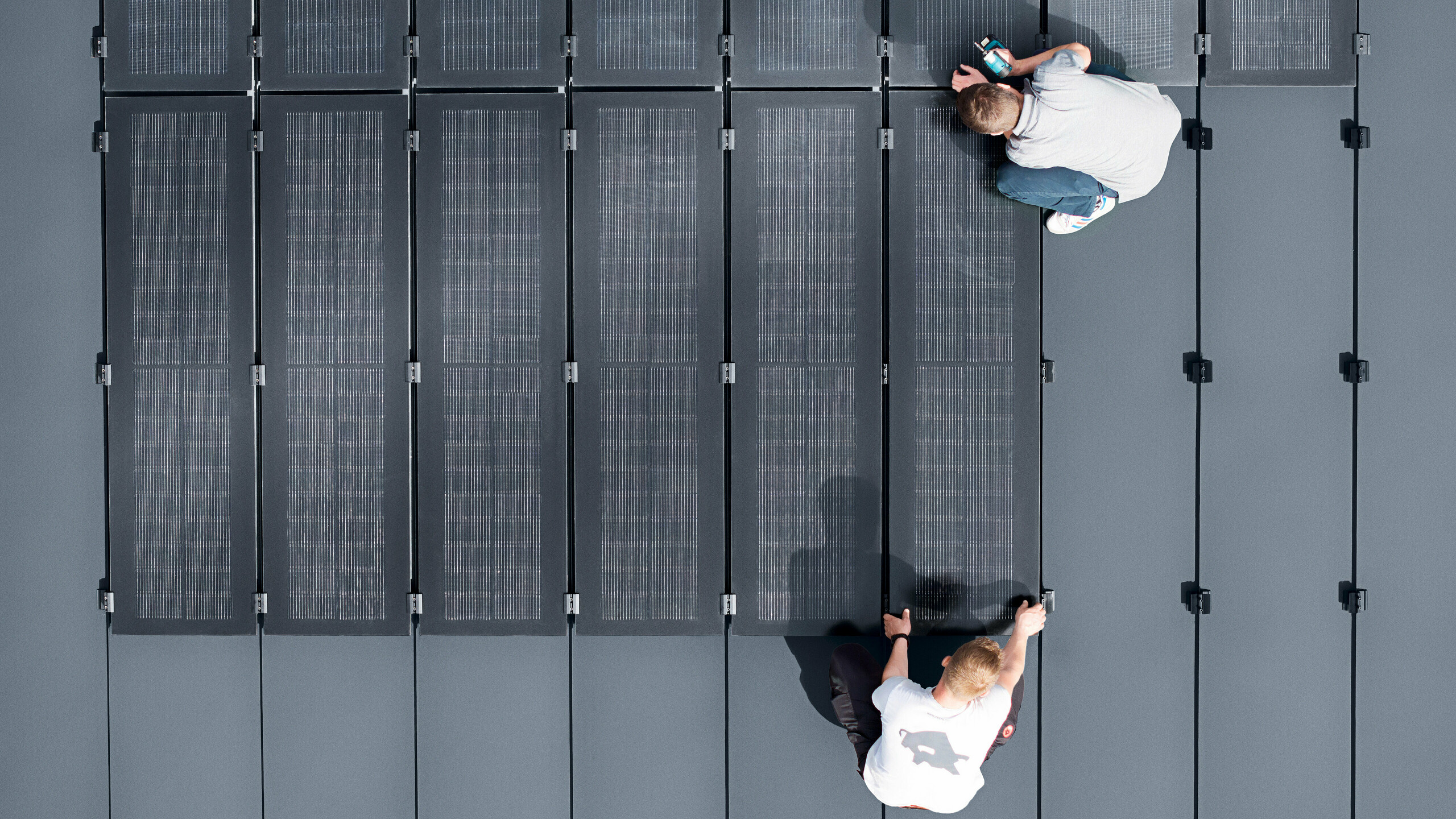 Zwei Personen installieren PREFALZ Solarmodule auf einer großen grauen Dachfläche, die mit Doppelstehfalzbahnen ausgelegt ist.