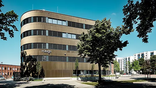 Dynamický architektonický pohled na policejní stanici 43 v Hamburku: Zakřivená novostavba s hliníkovou fasádou PREFA z extrudovaných profilů „Zacke“ ve střední bronzové barvě je v městské krajině výrazným poutavým prvkem. Jemně texturovaný povrch vytváří mezi horizontálními okenními pásy zářivé světelné a stínové efekty a zdůrazňuje plynulé linie fasády. Hliník PREFA přesvědčuje vysokou odolností proti korozi, trvanlivostí a nízkými nároky na údržbu – ideální pro udržitelná, funkční a architektonicky precizní řešení fasád.
