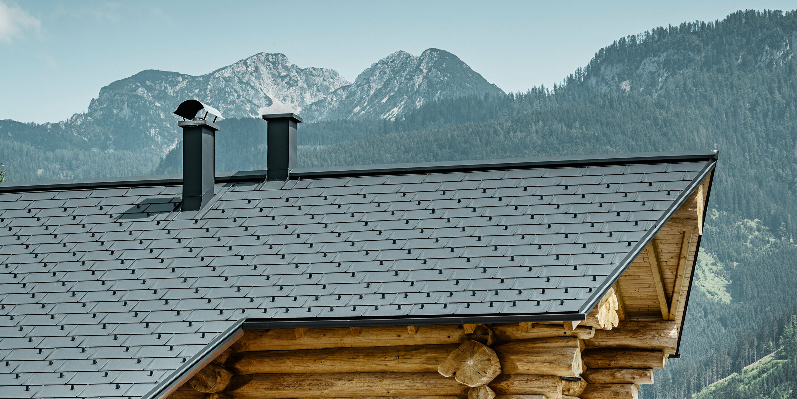 Působivý pohled na tradiční srub v rakouském Gosau proti působivému horskému panoramatu. Dům má hliníkovou střechu PREFA tvořenou střešními panely R.16 v barvě P.10 antracitové. Střecha v antracitové barvě vytváří moderní akcent rustikální dřevěné konstrukce. Vysoce kvalitní střešní panely dokonale zapadají do alpské krajiny, zatímco přirozenost dřevěného domu podtrhuje venkovský životní styl. Kombinace přírodních materiálů a estetického designu střechy dodává této nemovitosti nadčasové kouzlo.