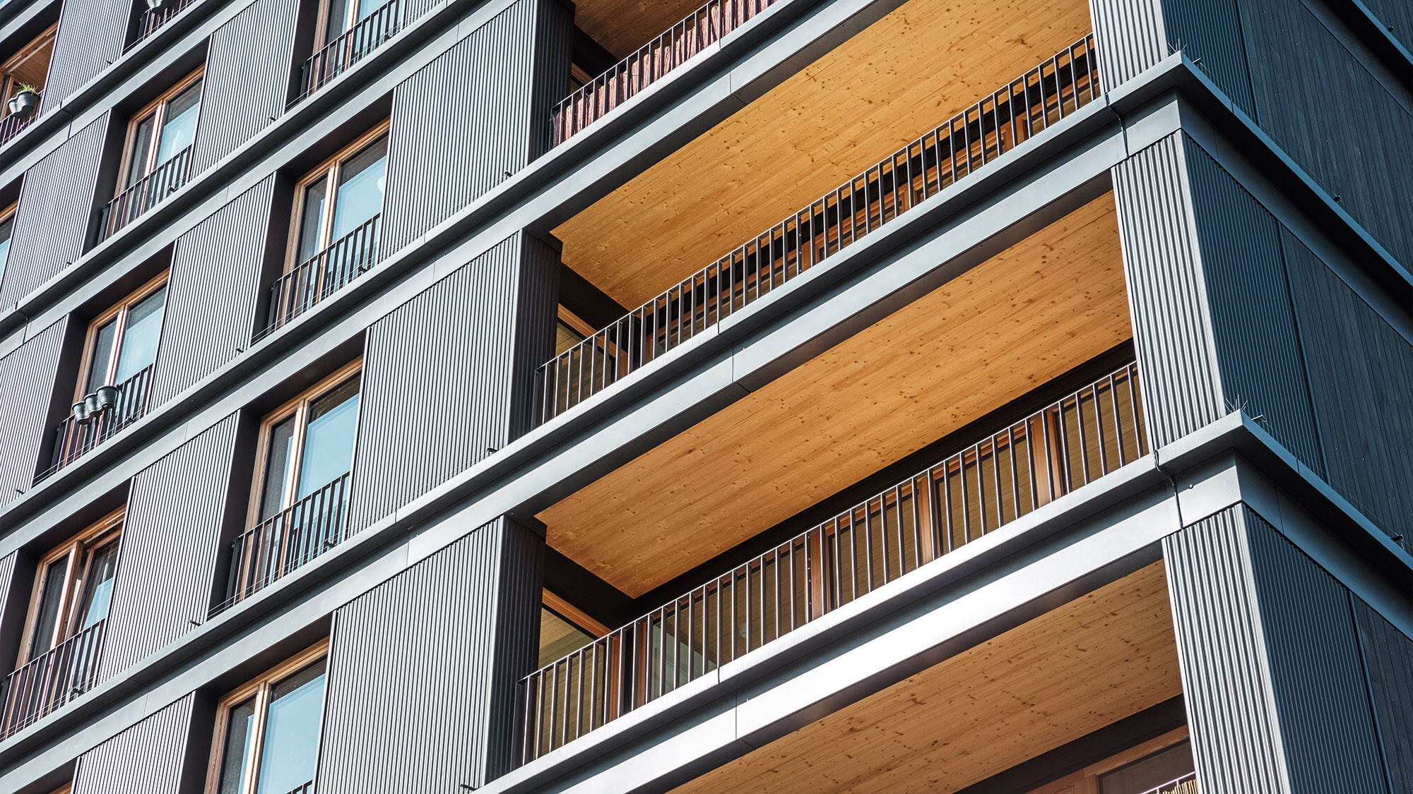 Mutlifunkční objekt z bočního pohledu zespodu: v pohledu hliníková profilová vlna, balkony a dřevem obložené balkonové stropy.