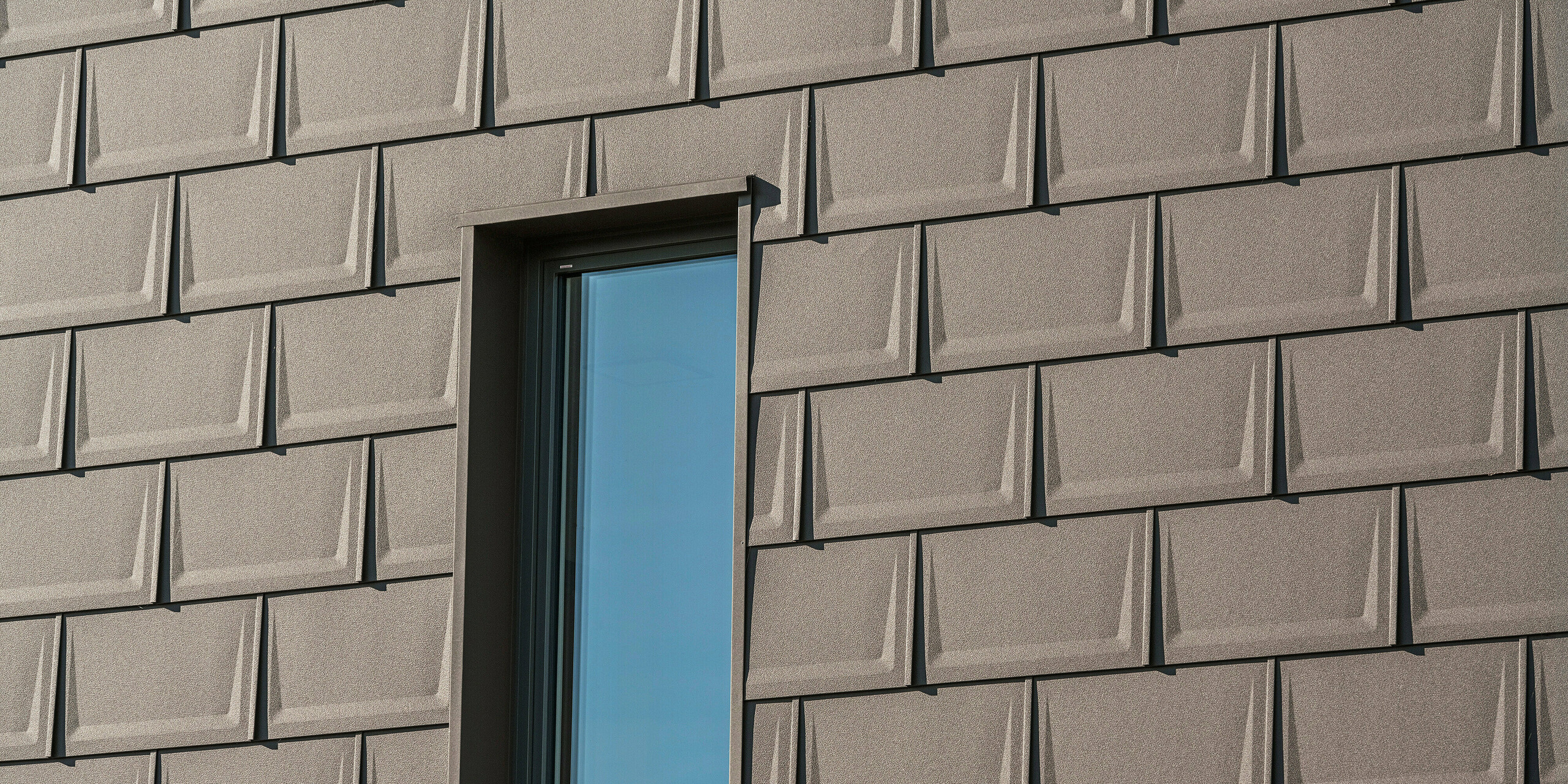Detailní záběr okna na fasádě rodinného domu v rakouském Neukirchenu, zvýrazněný střešním panelem PREFA R.16 v hnědé P.10. Jednotné pokládání panelů vytváří dynamickou texturu a dodává fasádě domu moderní ráz. Preciznost montáže a harmonické sladění barev podtrhují kvalitní konstrukční design, který stojí za hliníkovými výrobky PREFA.