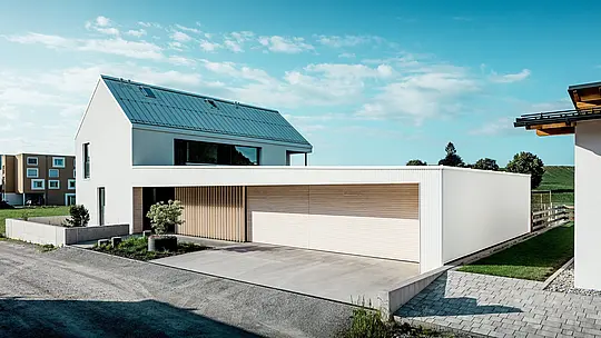 Obrázek ukazuje moderní rodinný dům ve Freistadtu, který se vyznačuje jednoduchým geometrickým tvarem a kombinací čistě bílé hliníkové fasády PREFALZ a dřevěného obložení. Dřevěný obklad garážových vrat vytváří teplý, přirozený kontrast a dokonale zapadá do moderního designu. Střecha, rovněž pokrytá PREFALZ, zajišťuje bezešvý a povětrnostním vlivům odolný povrch. Tato kombinace nenáročného hliníku a dřeva vytváří esteticky příjemné a funkční řešení pro udržitelnou architekturu. Prostorný, zelený venkovní prostor a jasná obloha v pozadí podtrhují moderní kvalitu života, kterou tento dům nabízí.