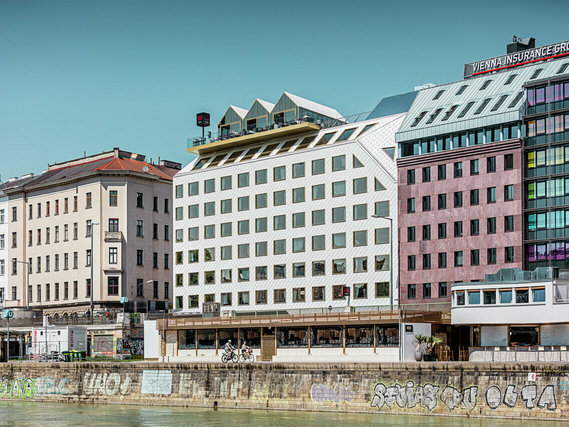 Pohled na břehy Dunajského kanálu ve Vídni s řadou budov různých architektonických stylů. V popředí je hotel "THE ROCK Radisson RED Vienna", rozpoznatelný podle charakteristicky bílé hliníkové fasády PREFA s falcovanými a fasádními šablonami 44 × 44 v barvě P.10 čistě bílé.