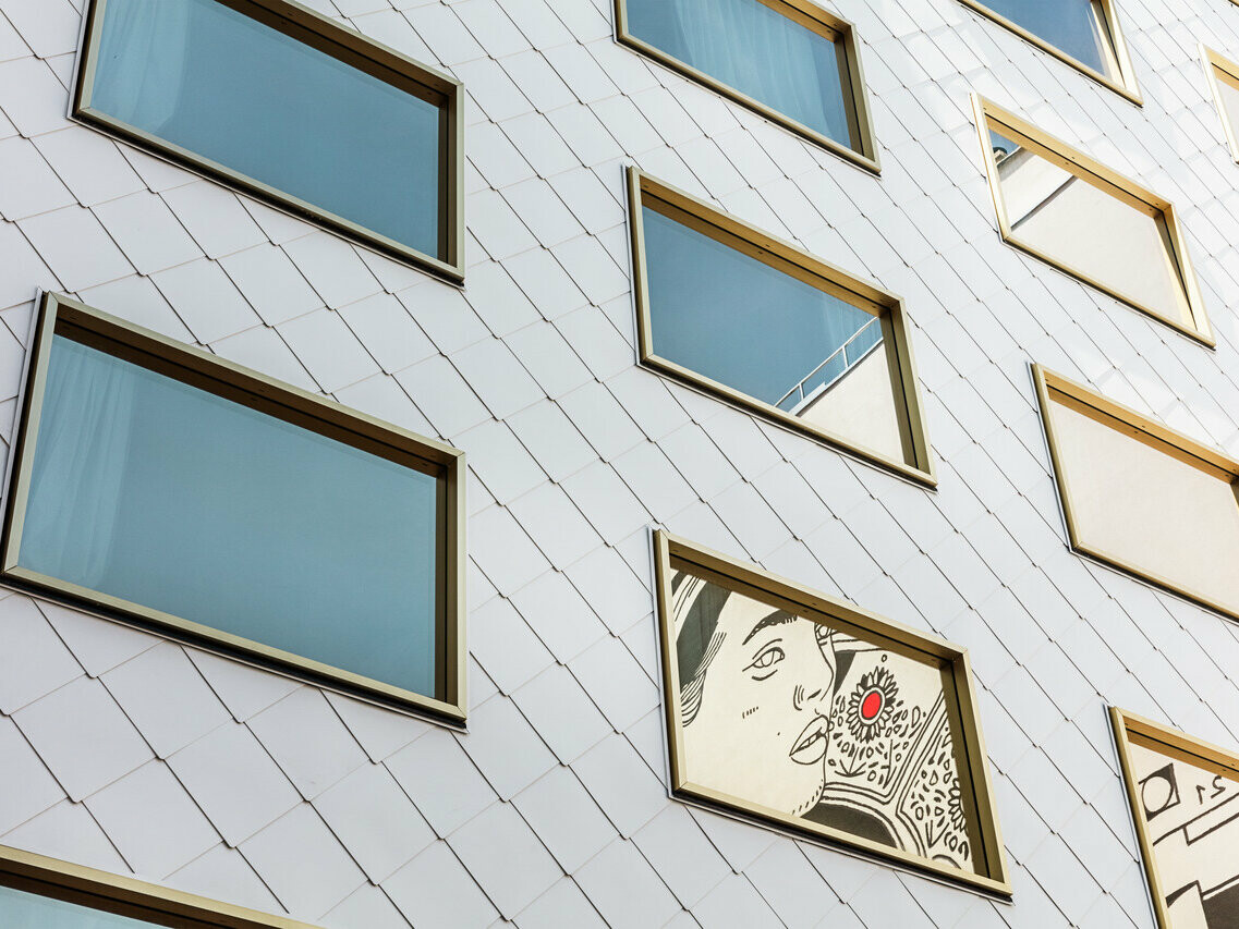 Detailní pohled na fasádu hotelu „THE ROCK Radisson RED Vienna“, která je obložena PREFA hliníkovými šablonami PREFA 44 × 44 v barvě P.10 čistě bílé. Mezi několika zrcadlově zasklenými okny se zlatými rámy vyniká okno, ve kterém je zasazeno umělecké dílo zobrazující portrét osoby s výrazným červeným akcentem.