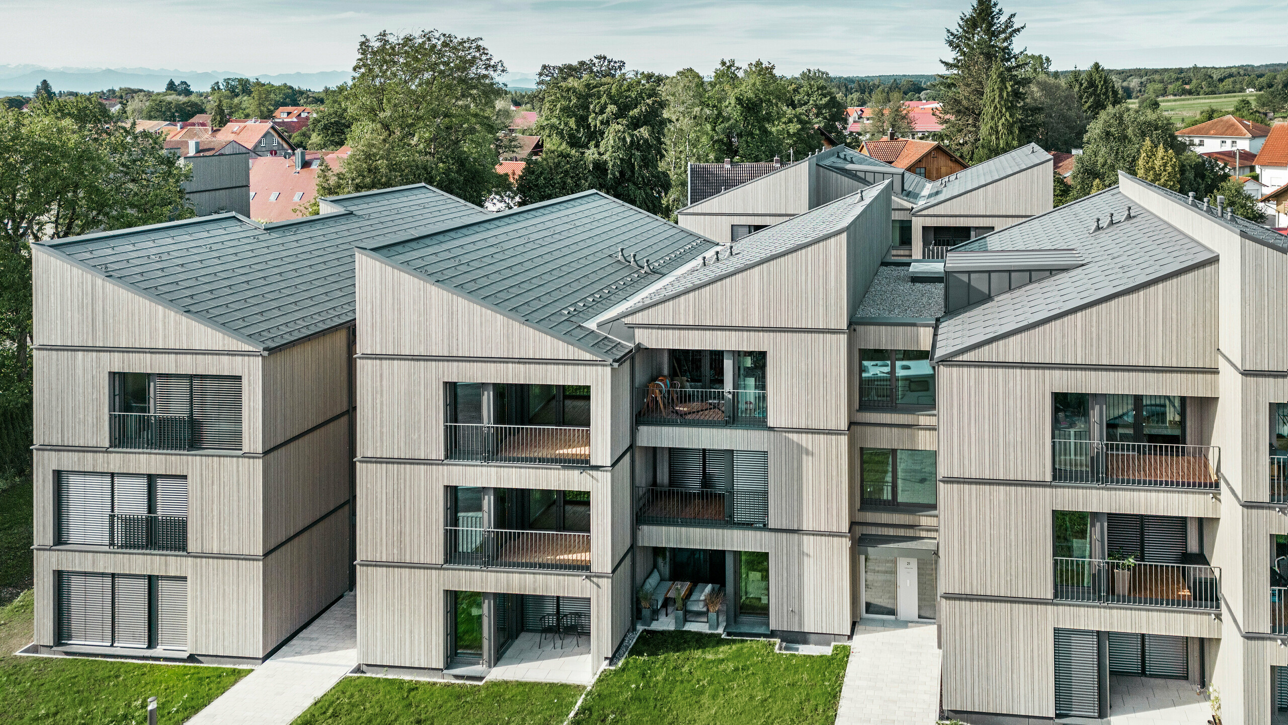 [Translate to Český:] Luftaufnahme des modernen Mehrfamilienhauses am Schmucker-Areal in Utting am Ammersee, mit nachhaltiger Holzhybridbauweise und eleganten Holzfassaden. Die Gebäude sind mit langlebigen PREFA Dachplatten R.16 in P.10 Dunkelgrau ausgestattet. Diese innovative Architektur, geplant von WWA Architekten aus München, zeichnet sich durch klare Linien und harmonische Integration in die grüne Umgebung aus. Die großzügigen Balkone und großen Fenster sorgen für eine helle und freundliche Wohnatmosphäre, während die hochwertigen Materialien für Langlebigkeit und Wetterbeständigkeit sorgen.