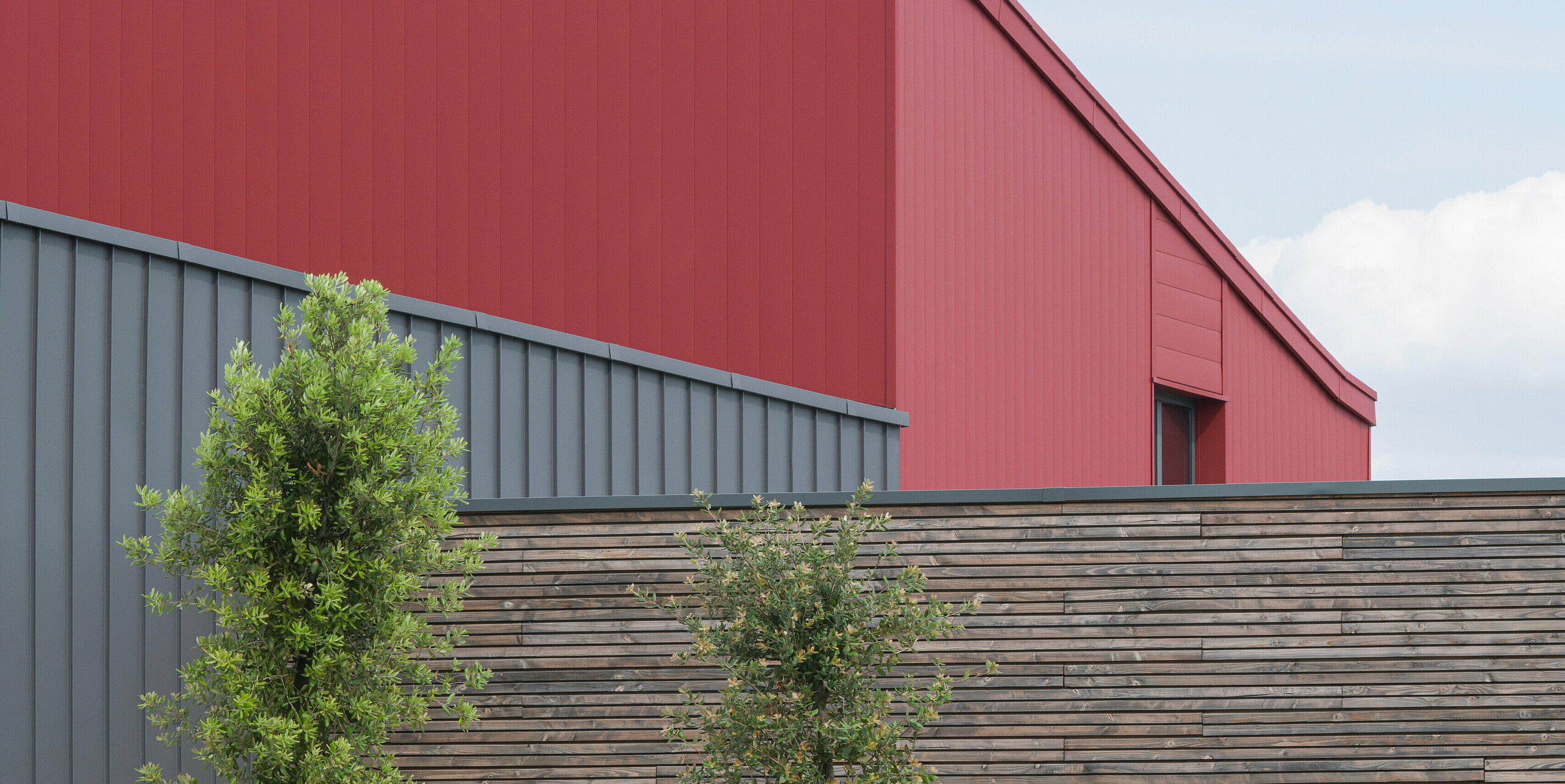 Moderní komerční budova s ​​nápadnou červenou hliníkovou fasádou. Výběr barvy hovoří jasnou architektonickou řečí. Strukturovaný, šedý hliníkový obklad tvoří jemný kontrast k výrazné červené.Odolný hliníkový obklad PREFALZ na fasádě a střeše poskytuje ideální ochranu před všemi povětrnostními vlivy. Doplňkový přírodní dřevěný obklad zprostředkovává teplo a přirozenost. Před budovou jsou vidět mladé stromky pečlivě zajištěné podpěrnými sloupky. Dobře udržovaný záhon dodává další barvu a život. Nabíjecí stanice pro elektromobily v popředí ukazuje závazek k udržitelným energetickým řešením.