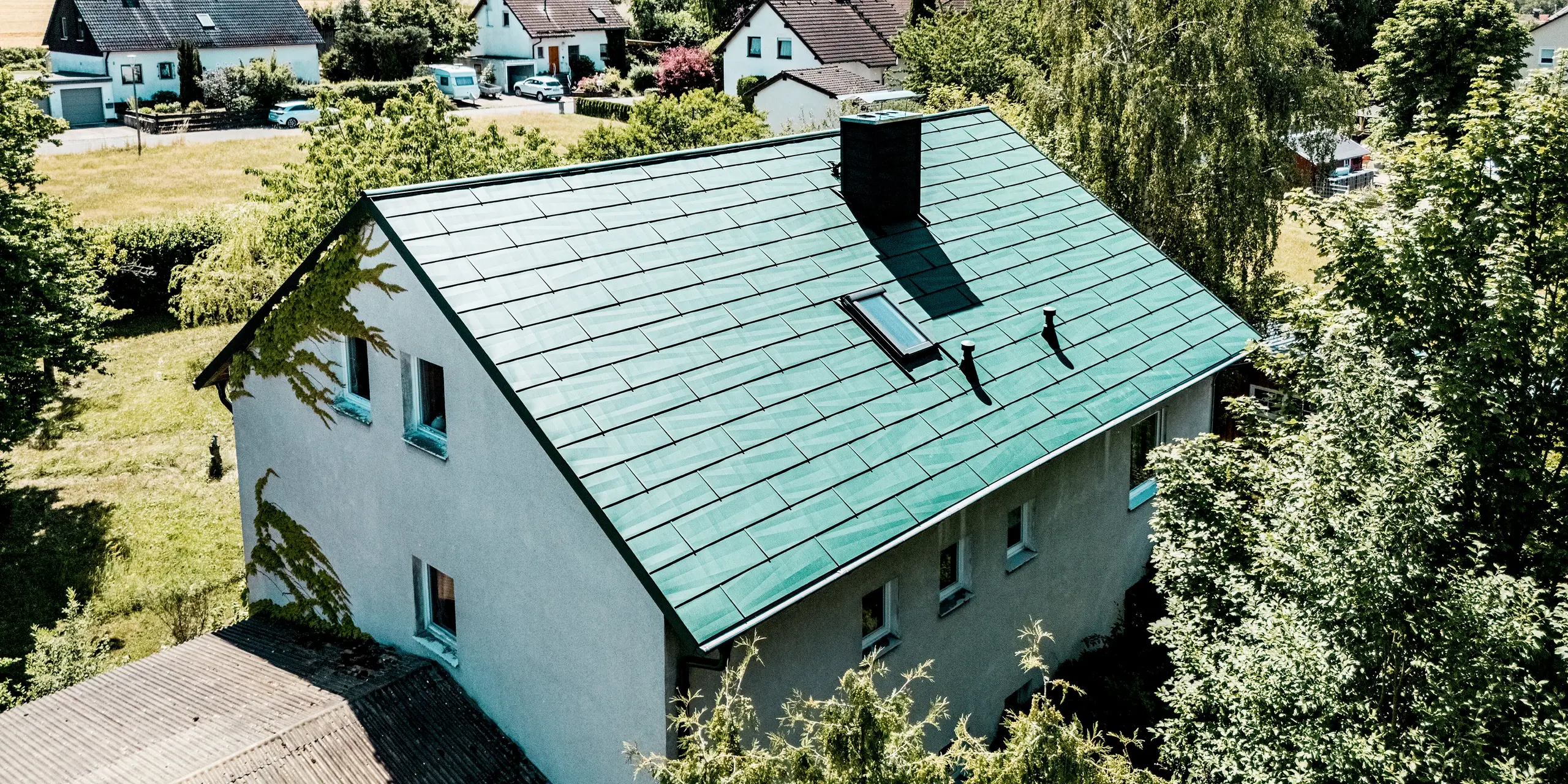 Einfamilienhaus in Schwarzenbach-Martinlamitz mit modernisiertem PREFA Dachpaneel FX.12 in P.10 Moosgrün. Die Aufnahme zeigt das sanierte Dach mit klarer Linienführung, markantem Dachfenster und elegant integriertem Kamin vor ländlicher Kulisse. Das Aluminiumdach besticht durch seine hochwertige Optik und die unregelmäßige Kantung der Paneele, die das Licht lebendig reflektiert. Wetterfest, langlebig und wartungsfrei bietet das PREFA Dach zuverlässigen Schutz und verleiht dem Haus eine moderne, charaktervolle Ausstrahlung mitten im Grünen.