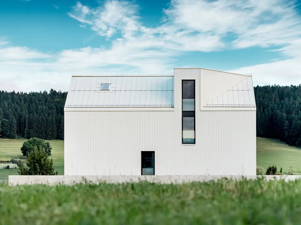 Obrázek ukazuje boční pohled na rodinný dům ve Freistadtu, který zdůrazňuje výraznou minimalistickou architekturu budovy. Čistě bílá hliníková fasáda PREFALZ a strmá sedlová střecha vytvářejí čistou, moderní siluetu, která harmonicky zapadá do zeleného okolí. Velká okna a elegantní vikýř zdůrazňují funkční design, zatímco vysoce kvalitní hliník zajišťuje estetickou trvanlivost i odolnost vůči povětrnostním vlivům. Díky svému jednoduchému tvaru a použití trvanlivých materiálů budova vyzařuje nadčasovou eleganci a představuje dokonalou kombinaci architektury a spojení s přírodou.
