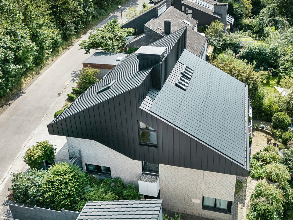 Schrägansicht eines modernen Einfamilienhauses in Meschede, Deutschland. Das Satteldach ist mit PREFALZ P.10 Schwarz gedeckt. Das Aluminiumdach setzt einen optischen Akzent und bietet dem Gebäude langfristigen Schutz. Die präzise Doppeldachkonstruktion erstreckt sich vom Dach über die Fassade bis zur Vorderseite. Umgeben von üppigem Grün und einer ruhigen Straße fügt sich das energieeffiziente Haus mit innovativen PREFALZ-Solarmodulen harmonisch in die natürliche Umgebung ein.
