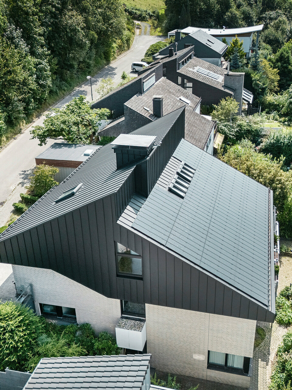 Schrägansicht auf ein modernes Einfamilienhaus in Meschede, Deutschland. Das versetzte Pultdach wurde mit PREFALZ in P.10 Schwarz verkleidet. Das Aluminiumdach dient als optisches Highlight und bietet dem Gebäude dauerhaften Schutz. Die präzise Doppelstehfalzdeckung zieht sich vom Dach über die Fassade an den Giebelseiten. Umgeben von üppigem Grün und einer ruhigen Straße fügt sich das energieeffiziente Gebäude mit den innovativen PREFALZ Solarmodulen perfekt in das naturnahe Wohnumfeld ein.