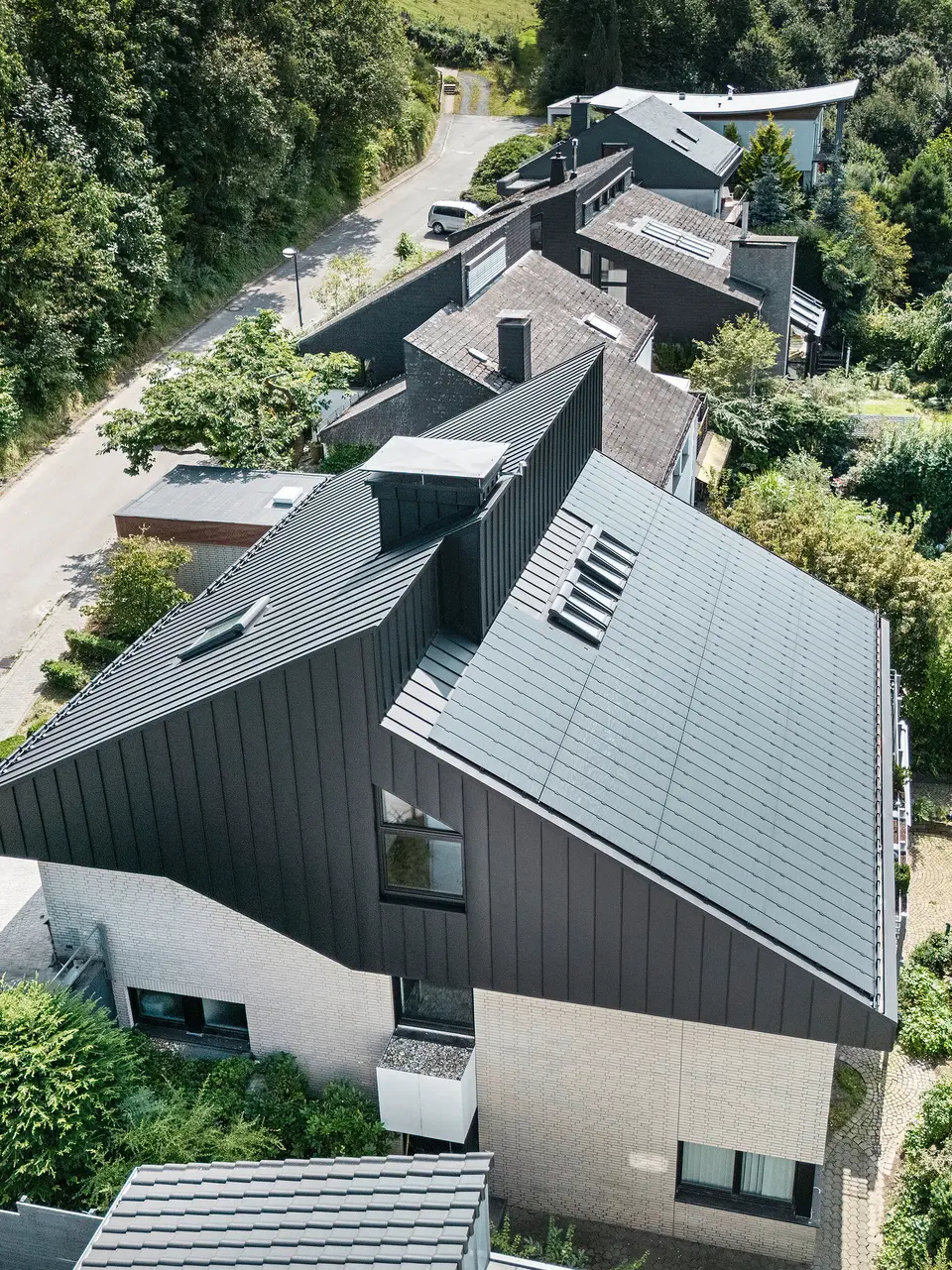 Schrägansicht eines modernen Einfamilienhauses in Meschede, Deutschland. Das Satteldach ist mit PREFALZ P.10 Schwarz gedeckt. Das Aluminiumdach setzt einen optischen Akzent und bietet dem Gebäude langfristigen Schutz. Die präzise Doppeldachkonstruktion erstreckt sich vom Dach über die Fassade bis zur Vorderseite. Umgeben von üppigem Grün und einer ruhigen Straße fügt sich das energieeffiziente Haus mit innovativen PREFALZ-Solarmodulen harmonisch in die natürliche Umgebung ein.