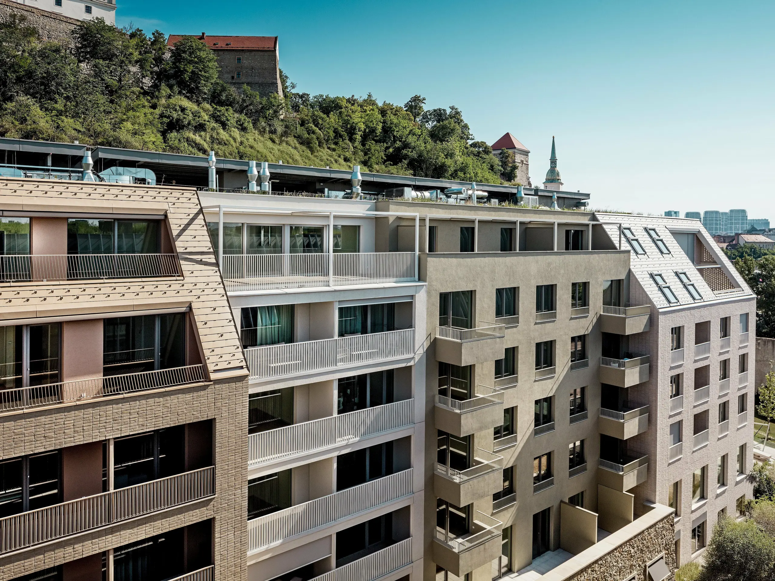 Městský rezidenční komplex v bratislavské čtvrti Vydrica, který se vyznačuje působivou souhrou moderní architektury a historického prostředí v blízkosti hradu: Strmé, precizně navržené střechy s hliníkovými šindeli PREFA v pískově hnědé a bílé barvě s čistými liniemi, vikýři a prostornými střešními terasami jsou vizuálně nejvýraznějšími prvky. Tato vysoce kvalitní hliníková řešení umožňují složité tvary střech, nabízejí mimořádnou trvanlivost, odolnost vůči povětrnostním vlivům a nenáročnou údržbu a vytvářejí udržitelný a esteticky propracovaný plášť budovy v městském kontextu.