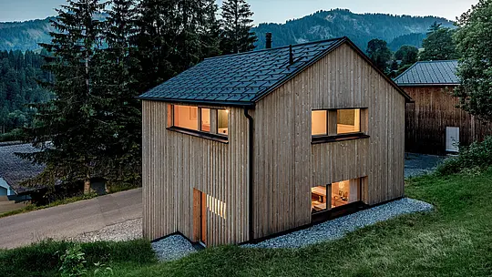 Z výšky se zdá, že Haus Immi je zasazen do horské krajiny Ebnitu. Teple osvětlená okna vytvářejí atmosférický kontrast s přírodní dřevěnou fasádou a černou solární střechou PREFA. Bezproblémově integrované solární střešní panely a střešní panely R.16 v černé barvě P.10 kombinují efektivní výrobu energie, vysokou odolnost a harmonický, moderní design střechy pro alpskou architekturu.