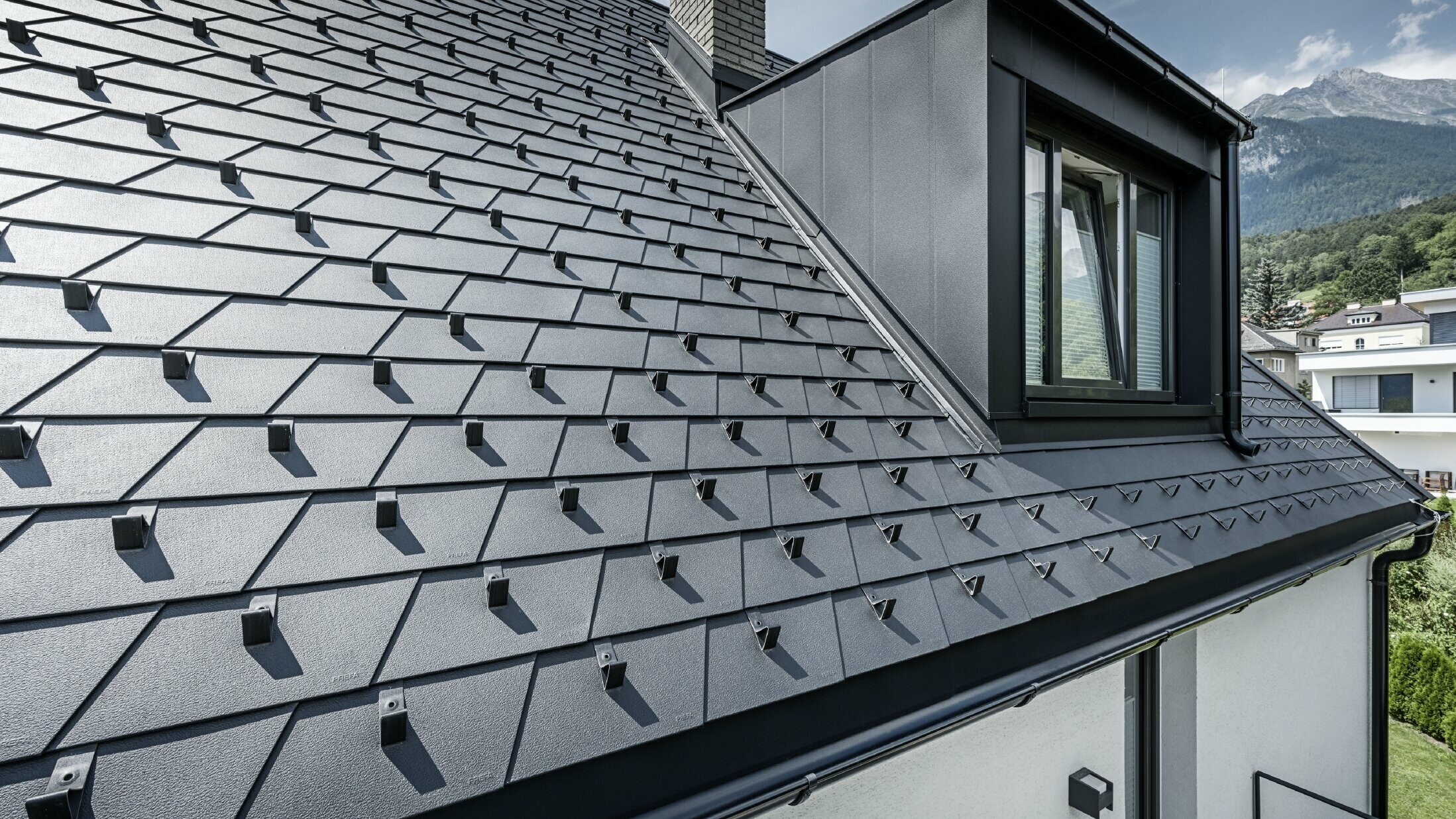 Detailní záběr střechy s vikýřem a oknem, na střešní ploše jsou položeny PREFA střešní šindele v černé barvě se sněhovými zábranami.