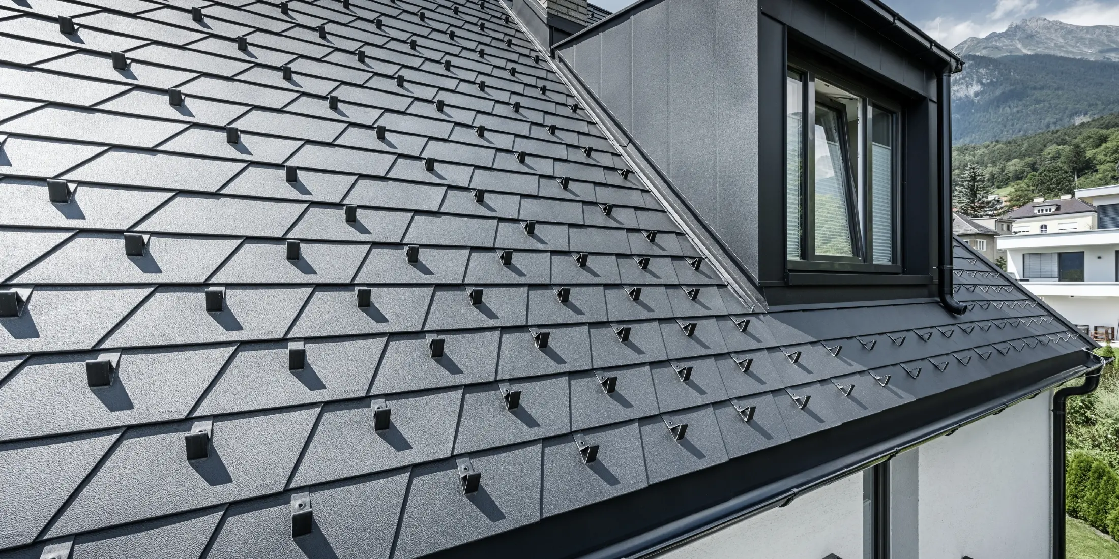 Detailní záběr střechy s vikýřem a oknem, na střešní ploše jsou položeny PREFA střešní šindele v černé barvě se sněhovými zábranami.